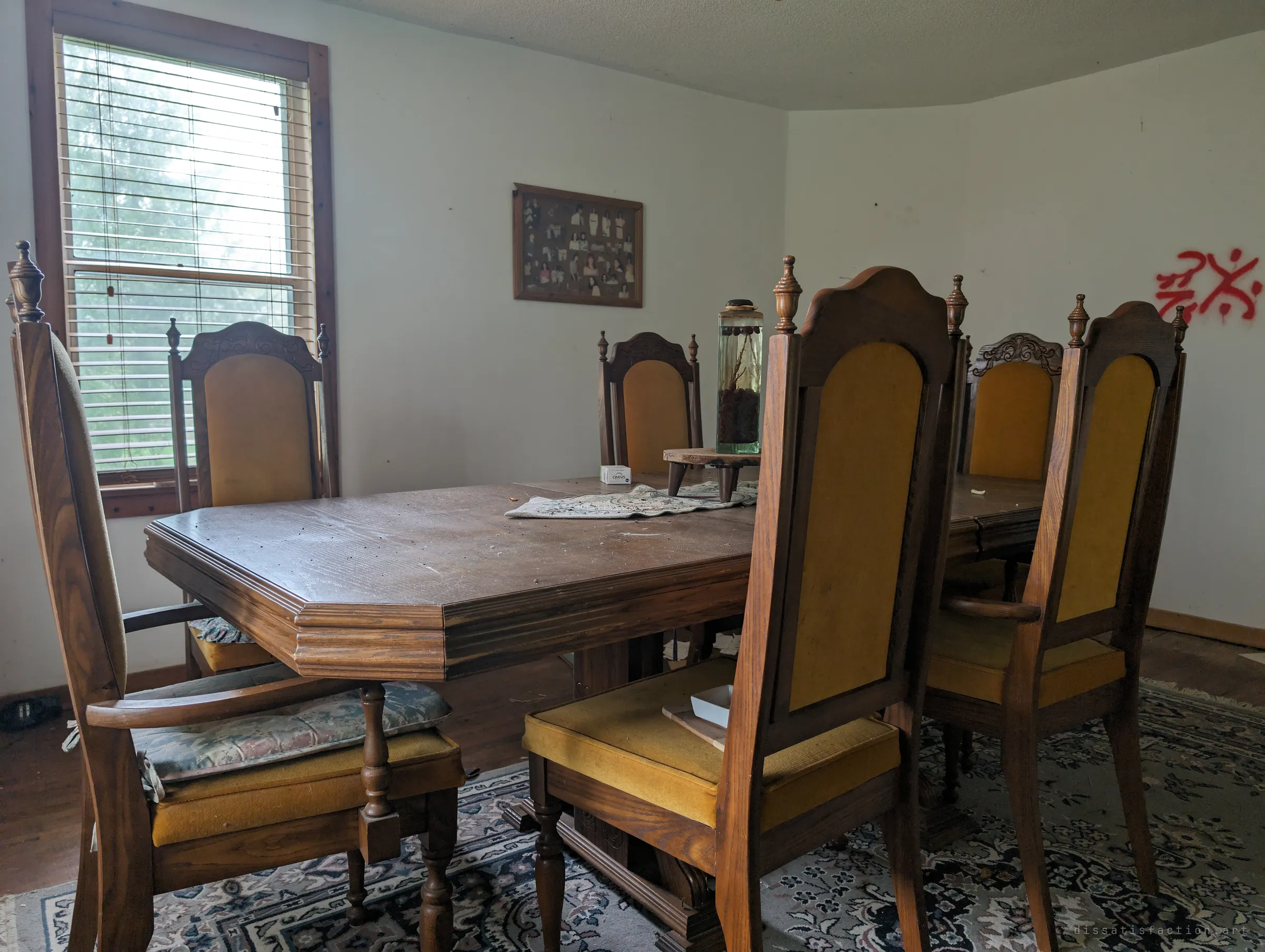 A formal dining room with carved high-backed chairs in mustard velvet around an octagonal table, a framed group portrait on the wall and a red graffiti tag in the corner. The set is still arranged for a dinner that never came.