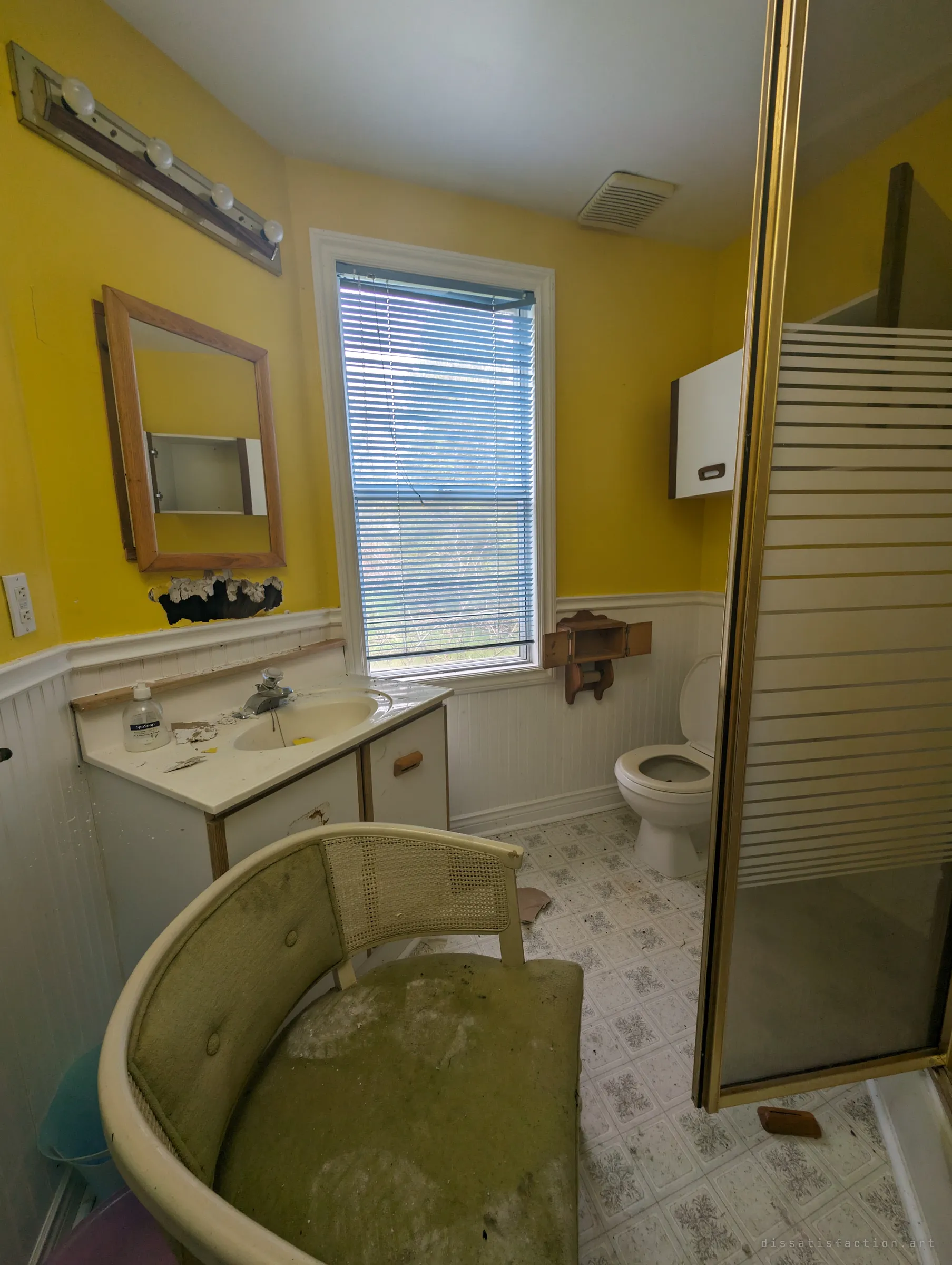 A small yellow bathroom with a vanity, a bare mirror cabinet and a sliding shower door, a mould-stained cane chair left in the middle of the floor. Daylight filters through the blinds onto the grime.