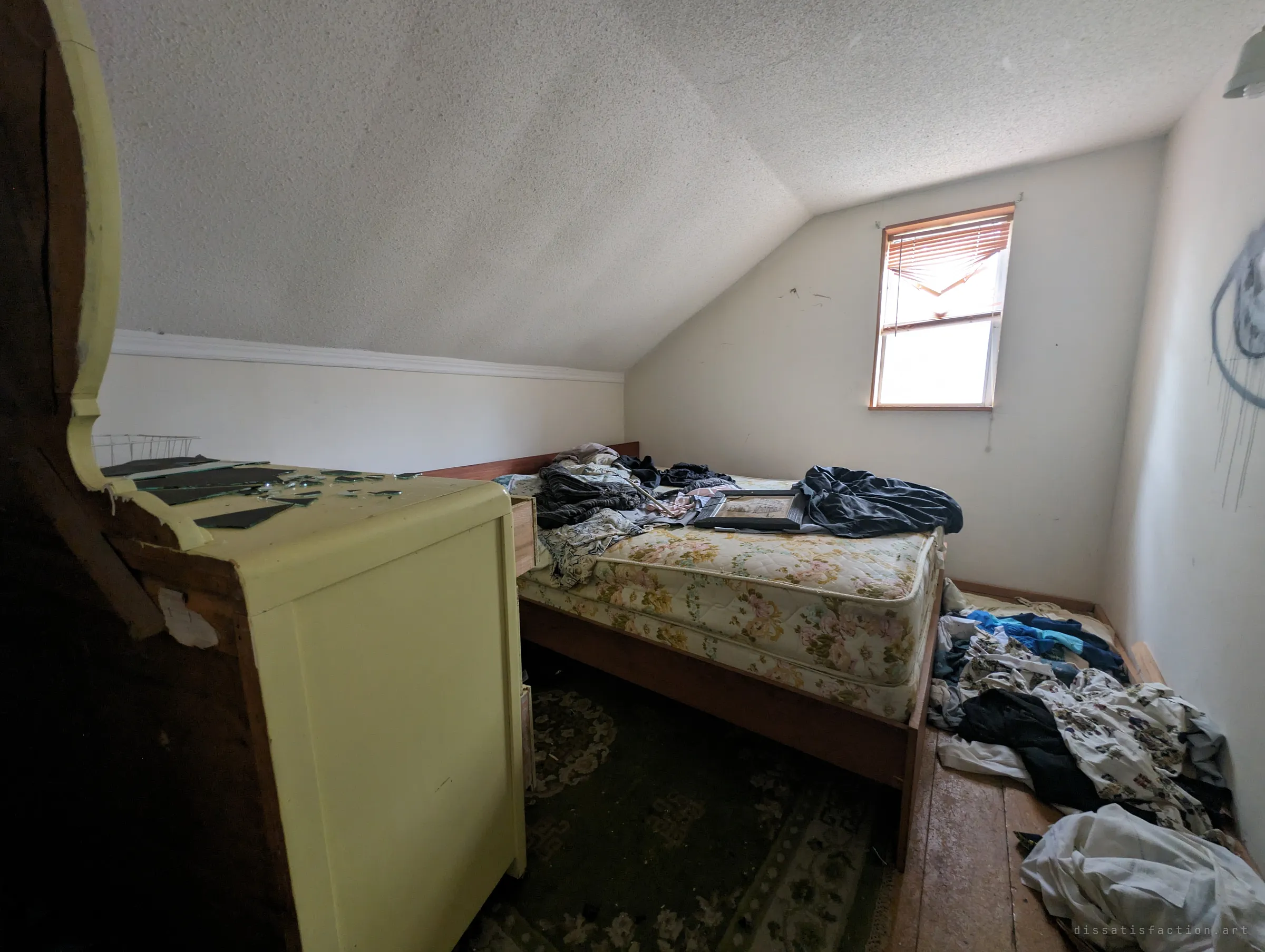 An attic bedroom under a sloped ceiling, with a bare floral mattress, clothes heaped on the floor and shards of a broken mirror on a pale yellow dresser. The upstairs rooms were left in disarray.