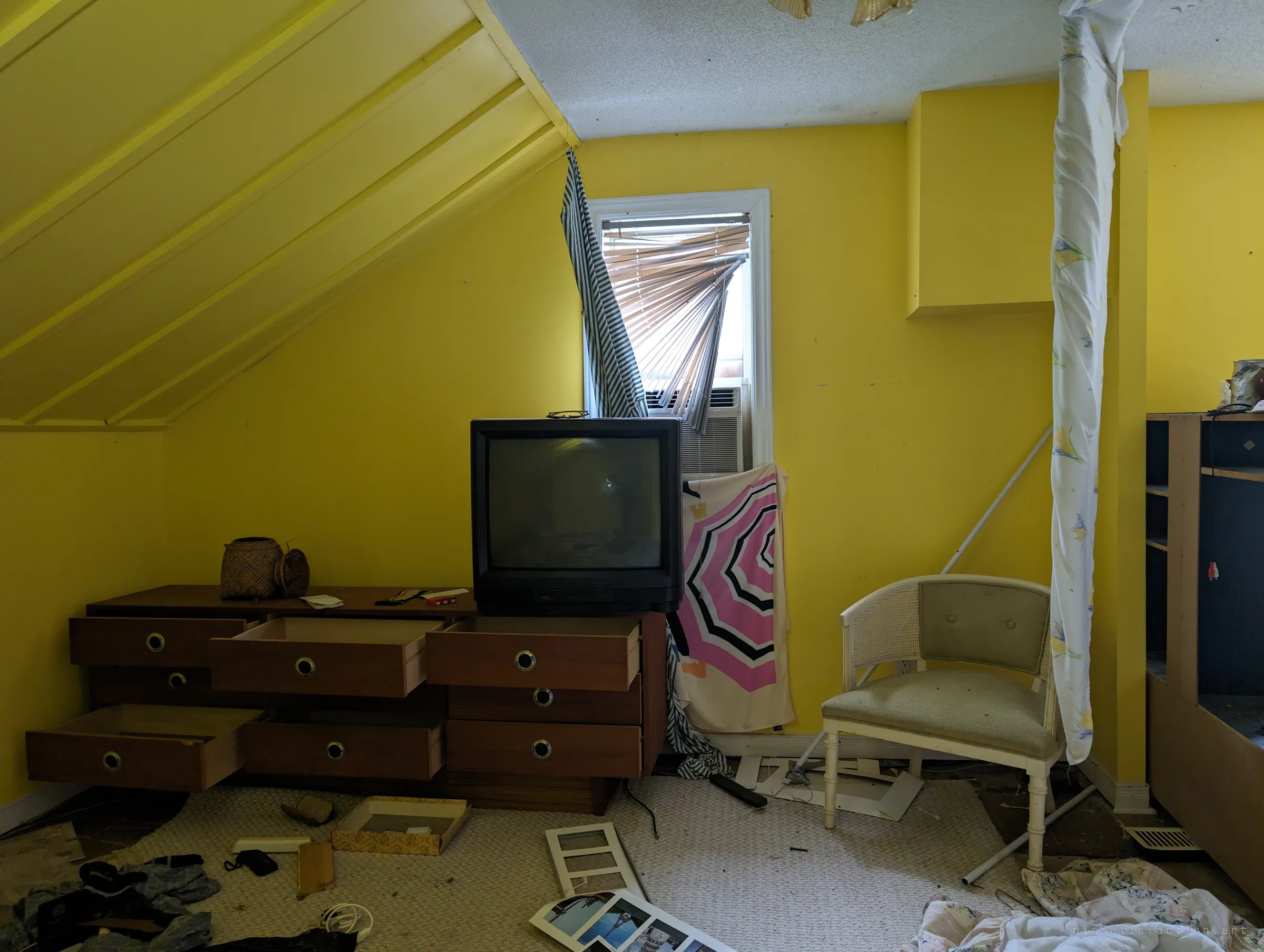 A yellow attic room with an old CRT television on a dresser of pulled-out drawers, a window air conditioner behind twisted blinds and scattered photos on the carpet. A time capsule of late-century furnishings.