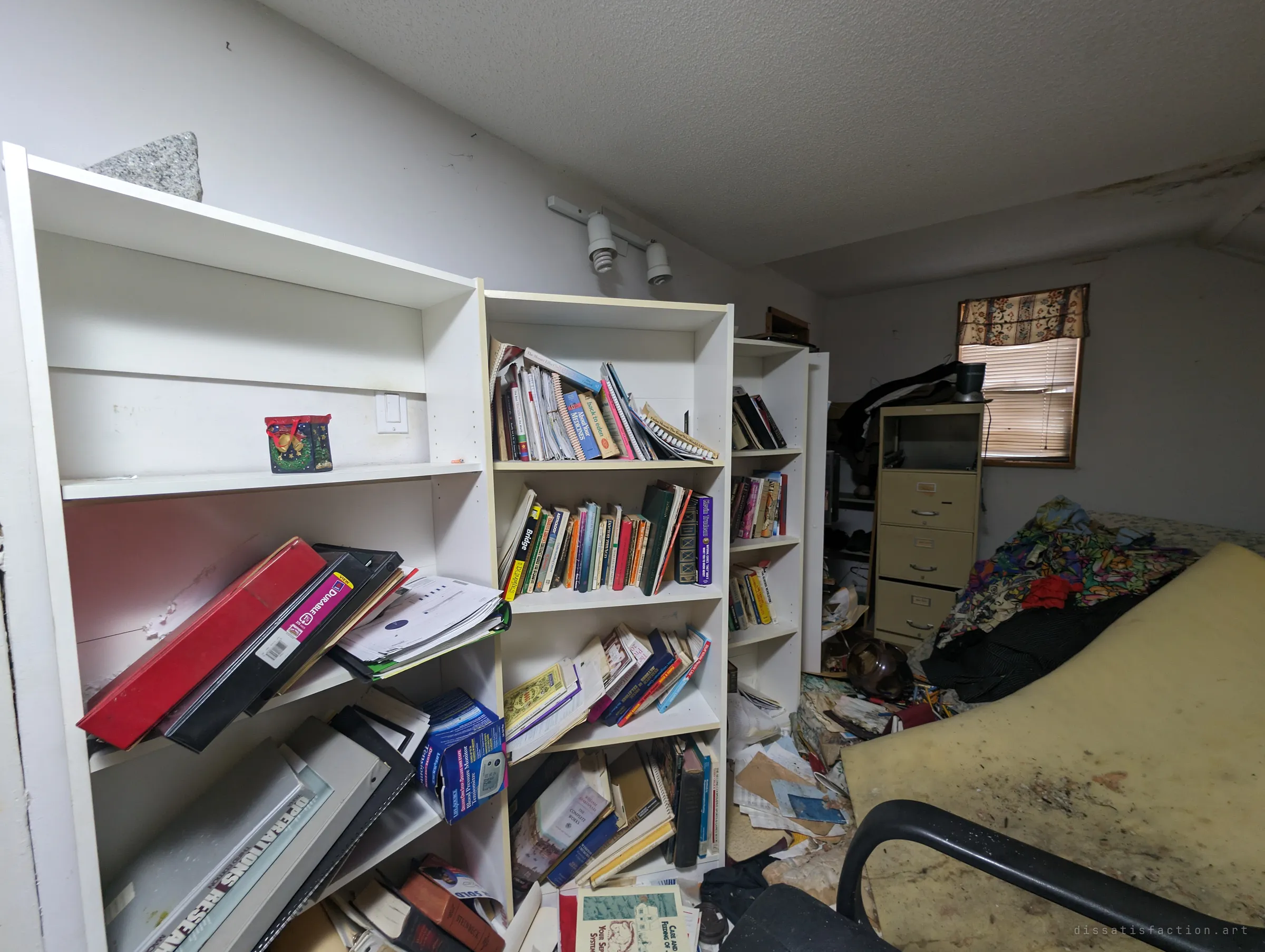 An attic study lined with white bookshelves, binders sliding off the shelves and papers spilling across the floor beside a stained foam mattress. A whole working life of books and files left behind.