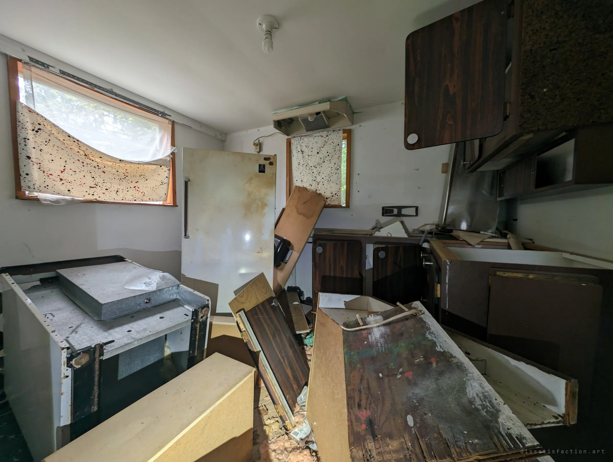 A wrecked kitchen with the range pulled out, cabinet doors torn off and a rusting upright fridge, windows covered with paint-splattered sheets. Daylight filters through the makeshift curtains onto the debris.