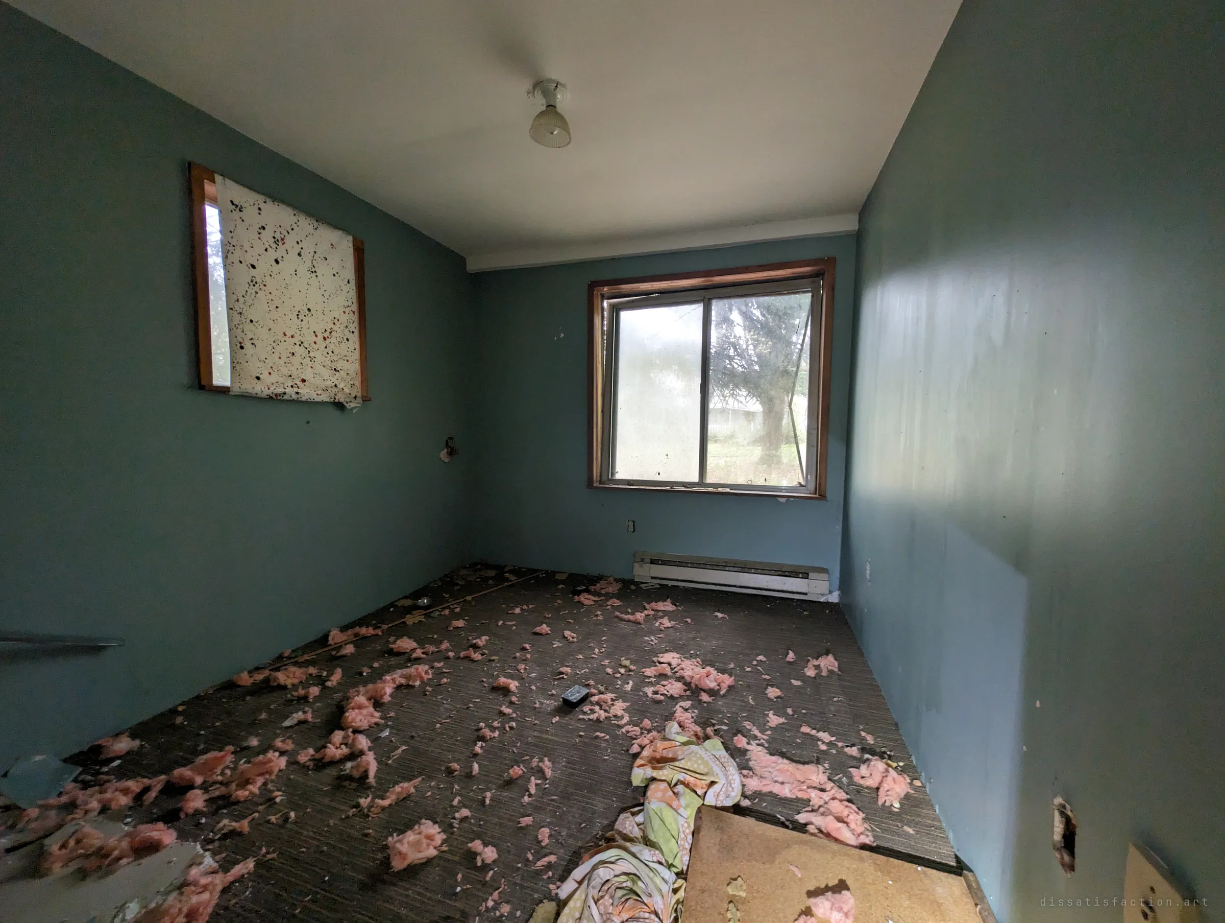 An empty teal bedroom with pink insulation scattered over the dark carpet and a splatter-painted sheet over one window. The quiet, stripped room contrasts with the cluttered spaces elsewhere in the house.