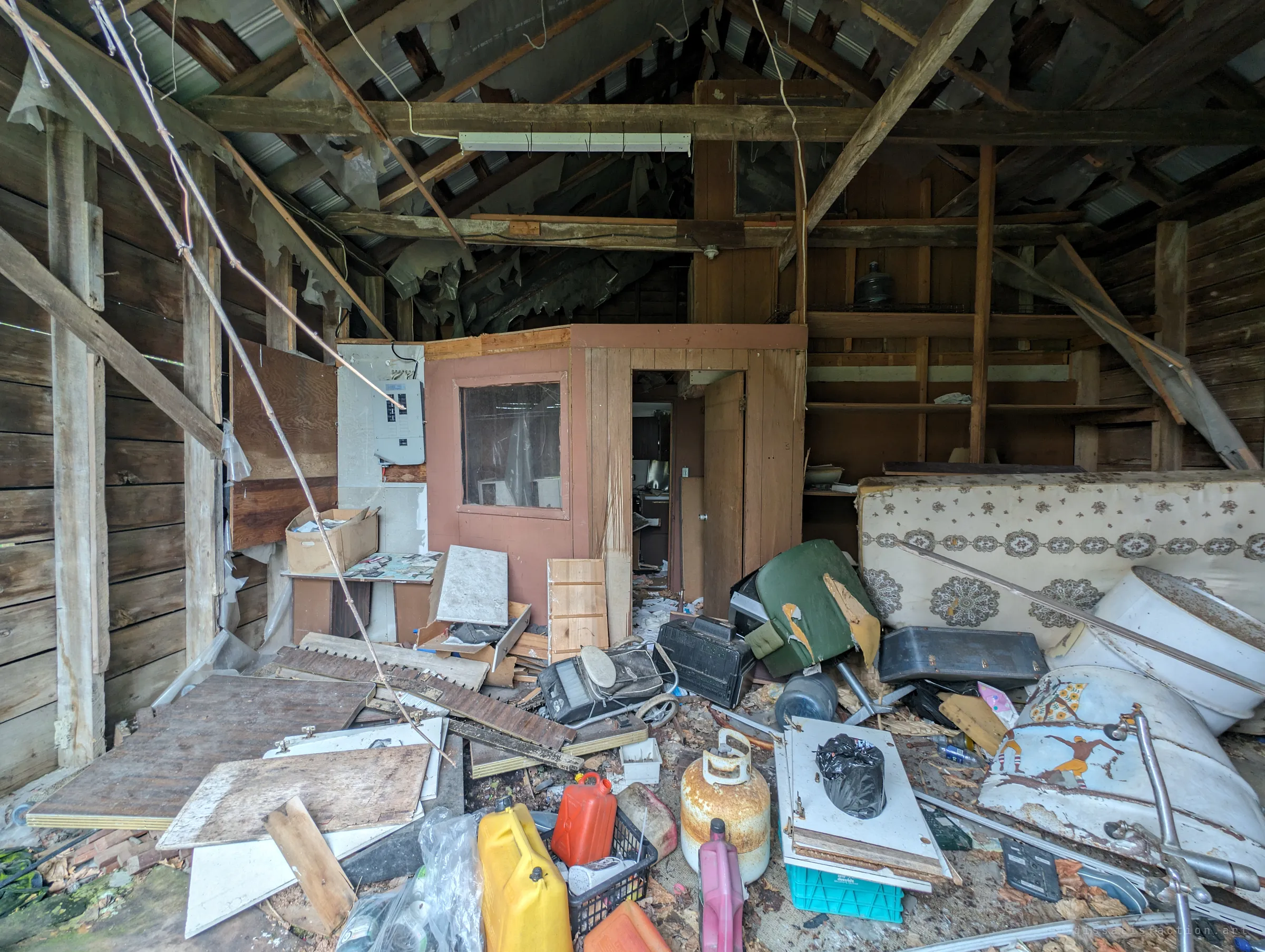 Inside a timber garage with a shredded roof liner hanging from the rafters, a small wood-panelled office booth built into the corner, and fuel cans, a propane tank and an old tub heaped in front. Layers of abandoned household life piled into one shed.
