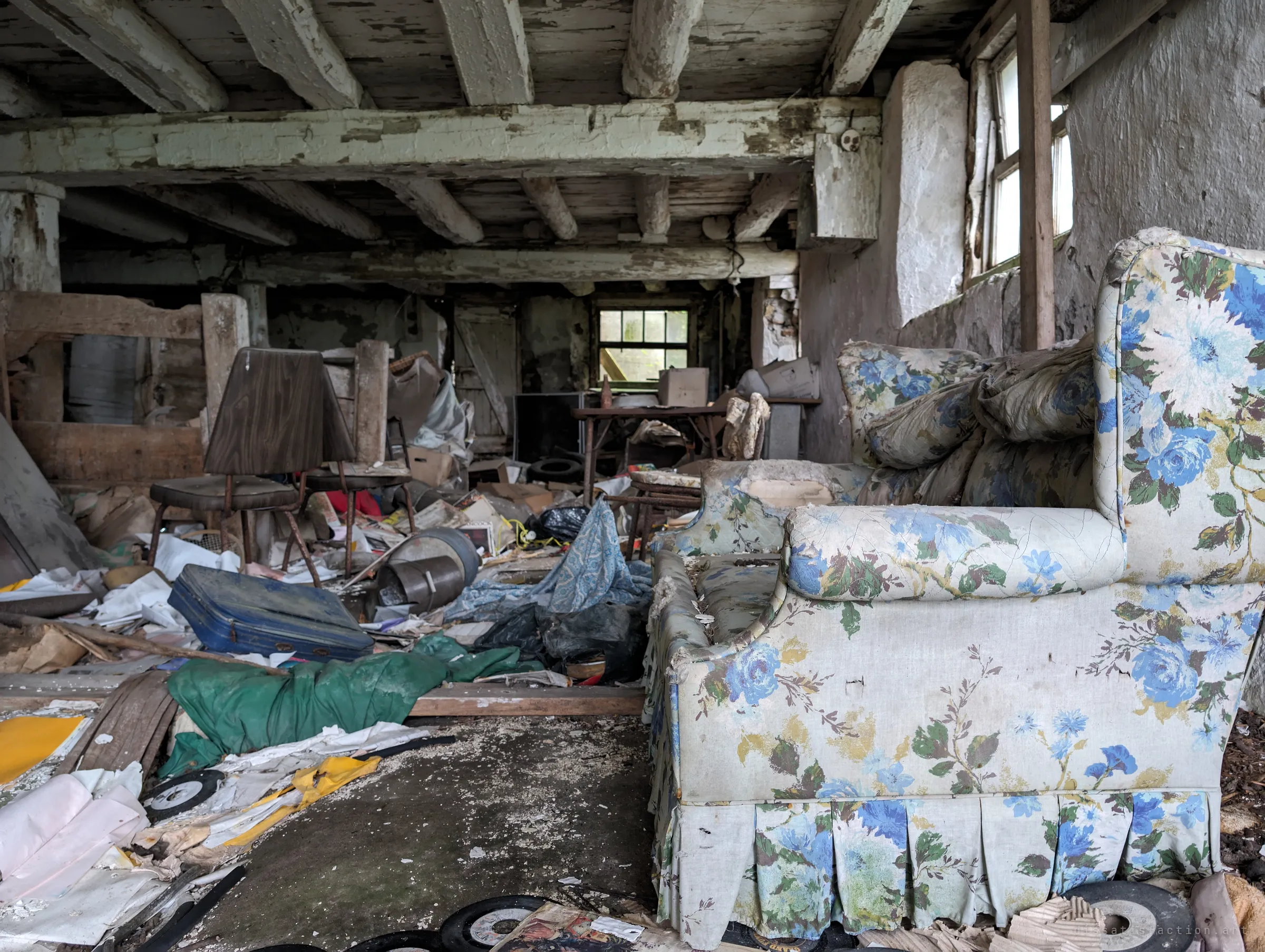A floral armchair sits in a whitewashed stone basement under heavy peeling beams, surrounded by chairs, a blue suitcase and scattered 45 records. The room feels frozen mid-clear-out.