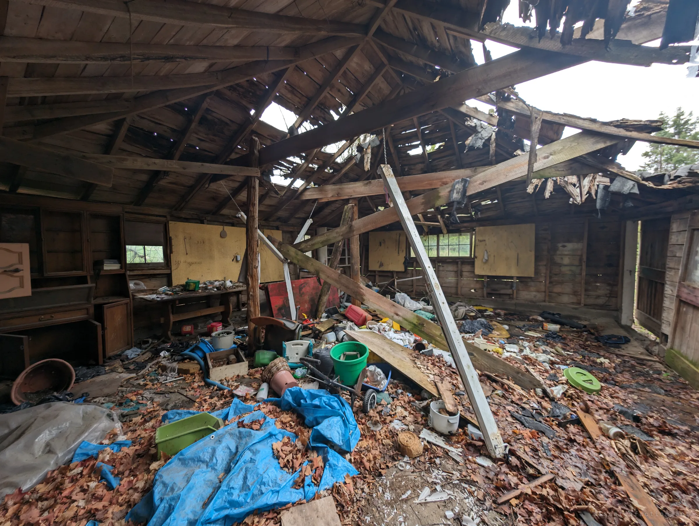 A workshop whose roof has partly collapsed, beams fallen at angles and sky showing through, the floor buried in autumn leaves, tarps and buckets. Pegboard walls still hold a few tools.