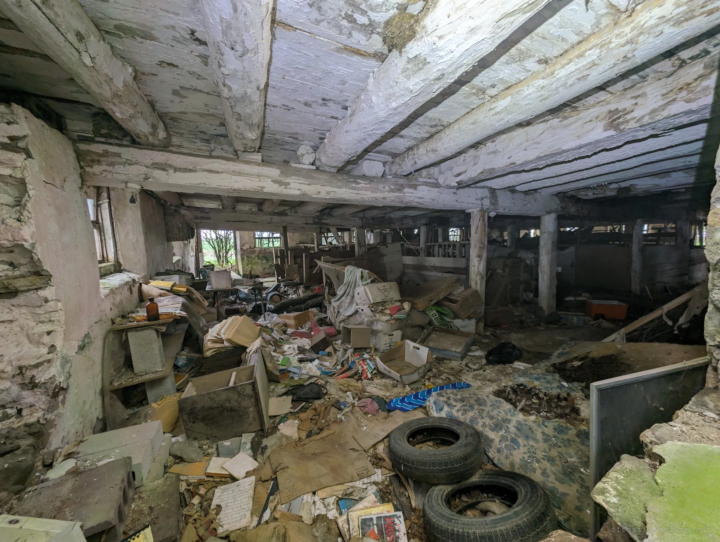 The lower level of an old stone barn, its low whitewashed beams peeling over a floor of boxes, papers, tyres and an old mattress. Rows of stall posts fade into the dark at the back.