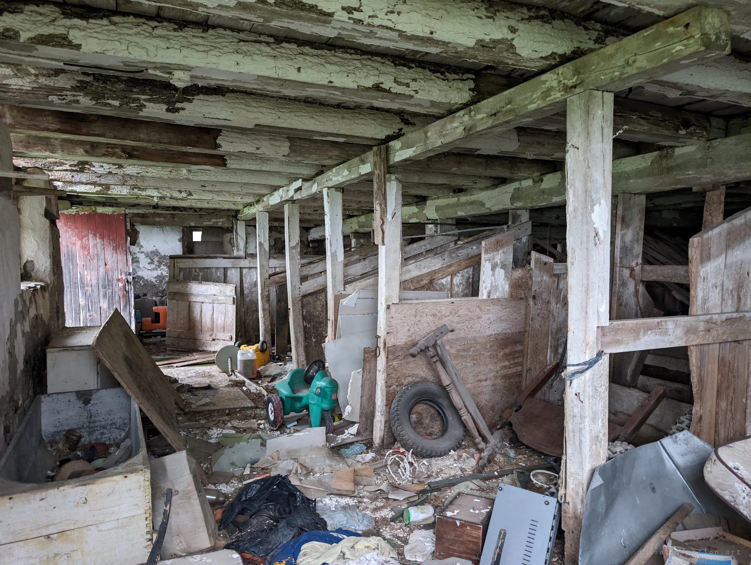 A row of wooden animal stalls under flaking green-white beams, a red plank door at the far end and a child's green ride-on toy among the junk. Farm use and family life overlap in the same space.