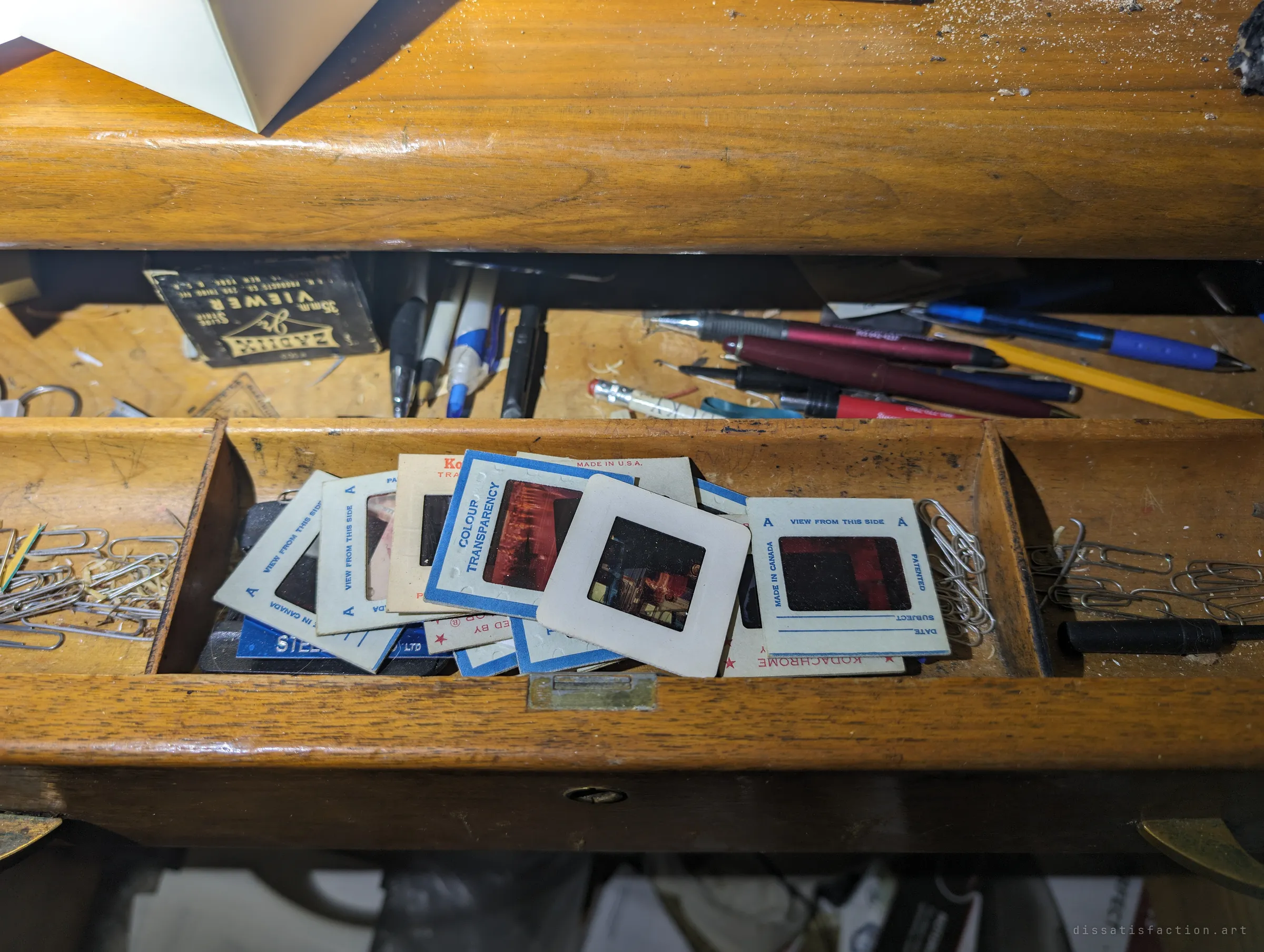 An open desk drawer full of old 35mm colour slides in paper mounts, with pens, paper clips and a slide viewer box behind. Someone's private photographs left in the dark.