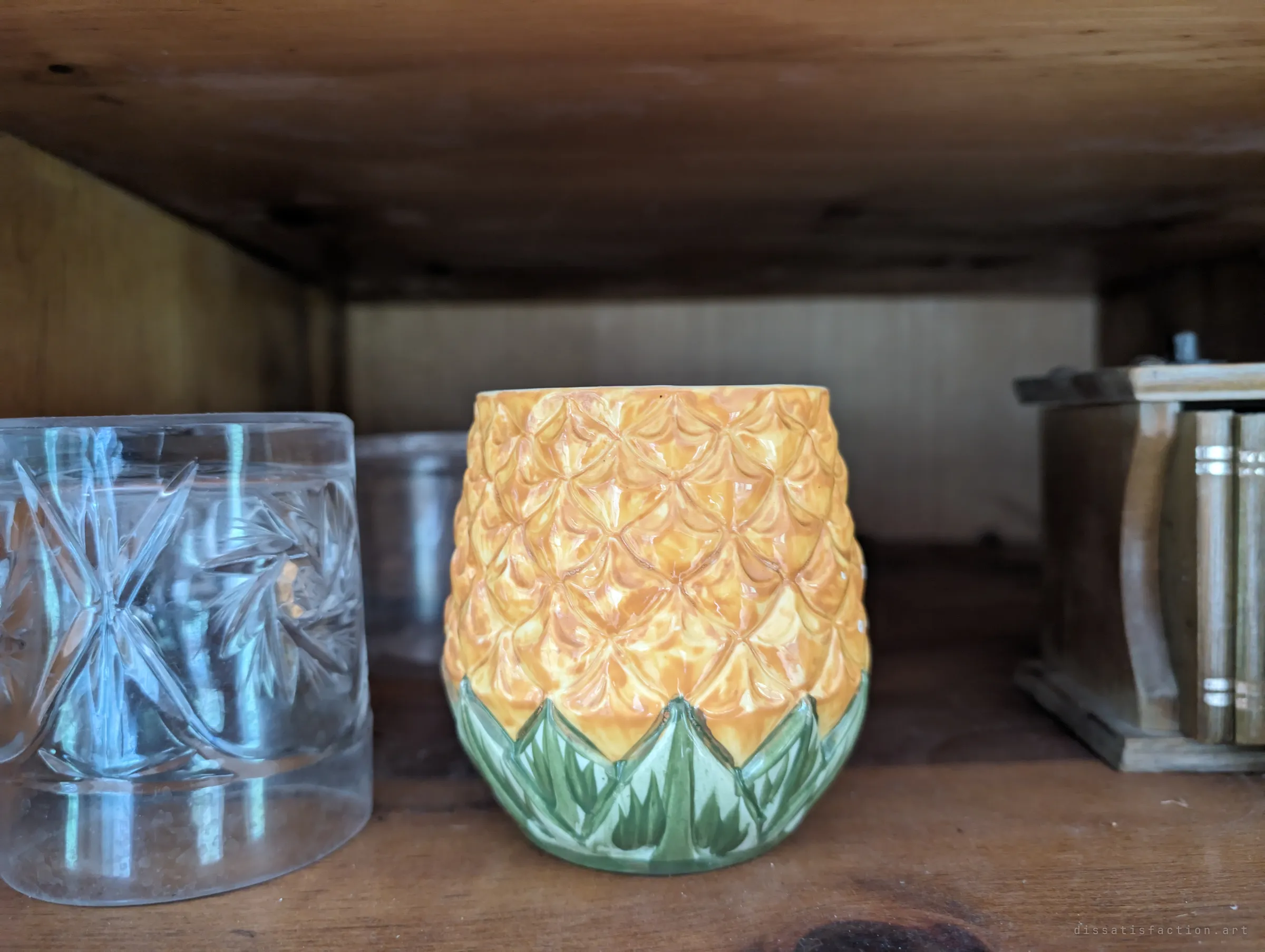 A glazed ceramic pineapple cup on a wooden shelf between a cut-glass tumbler and a small box of coasters. A small, cheerful detail that survived the decay.