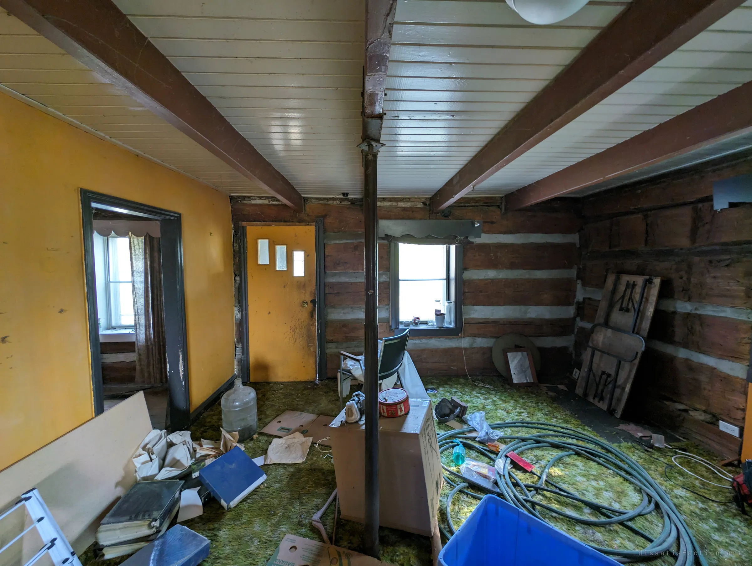 A low-beamed room with log walls, a yellow plaster wall and shaggy green carpet strewn with hoses, binders and cardboard. A steel post props up the sagging centre beam.