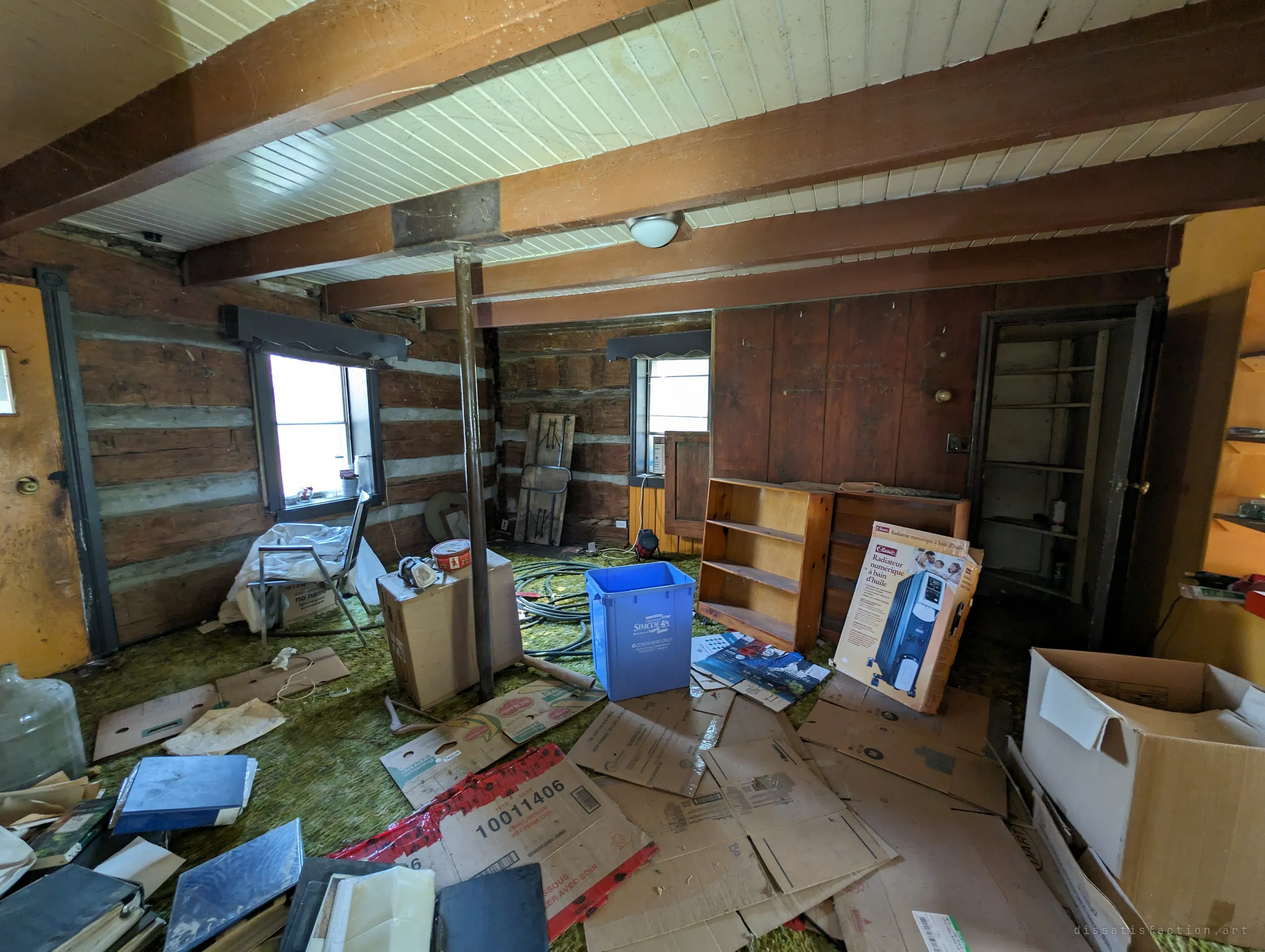 The same log-walled room seen from the other side: flattened boxes, a blue recycling bin, empty bookshelves and a boxed space heater left on the green carpet. It shows how the house was half packed and then abandoned.