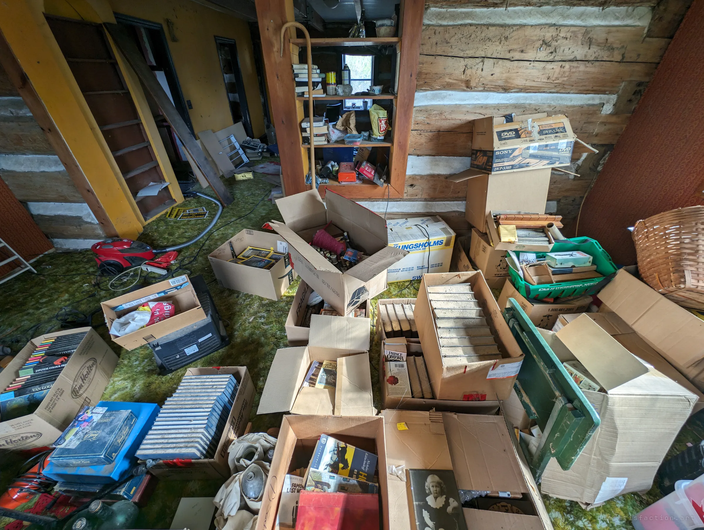 Dozens of open cardboard boxes of books, DVDs and keepsakes cover the floor in front of a built-in shelf and a steep wooden stair. An old framed portrait of a child sits among them.