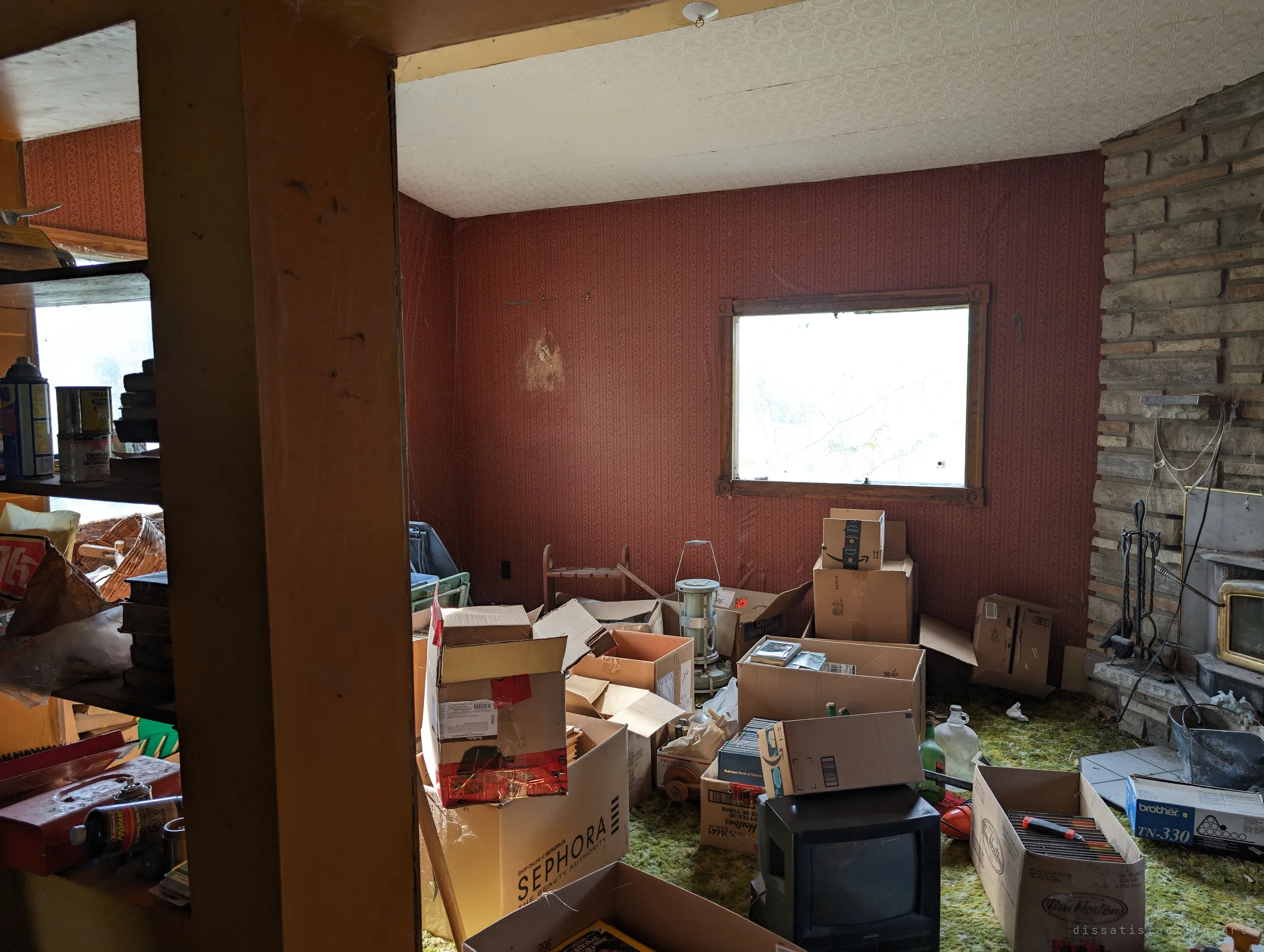 A red-wallpapered sitting room piled with boxes, an old kerosene heater and a small CRT television, beside a stone fireplace. Light from a bare window washes over the clutter.