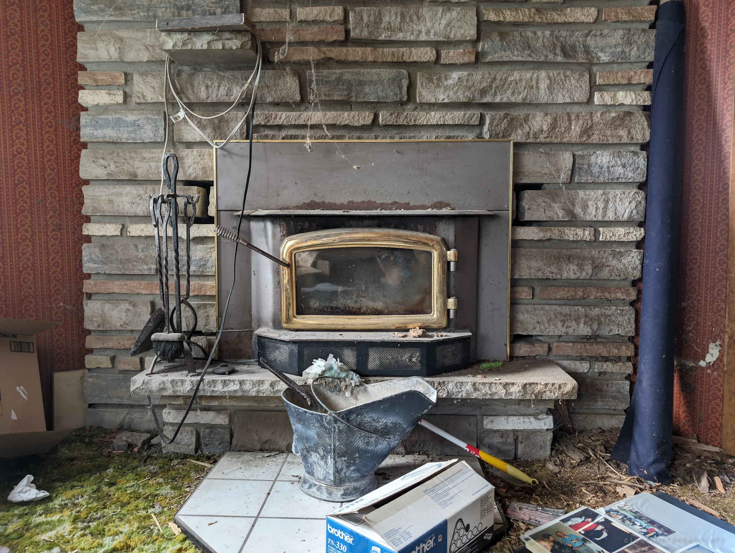 A fieldstone fireplace with a brass-doored insert, iron tools and a dusty coal scuttle; a family photo album lies open on the floor. The personal photos make the leaving feel sudden.