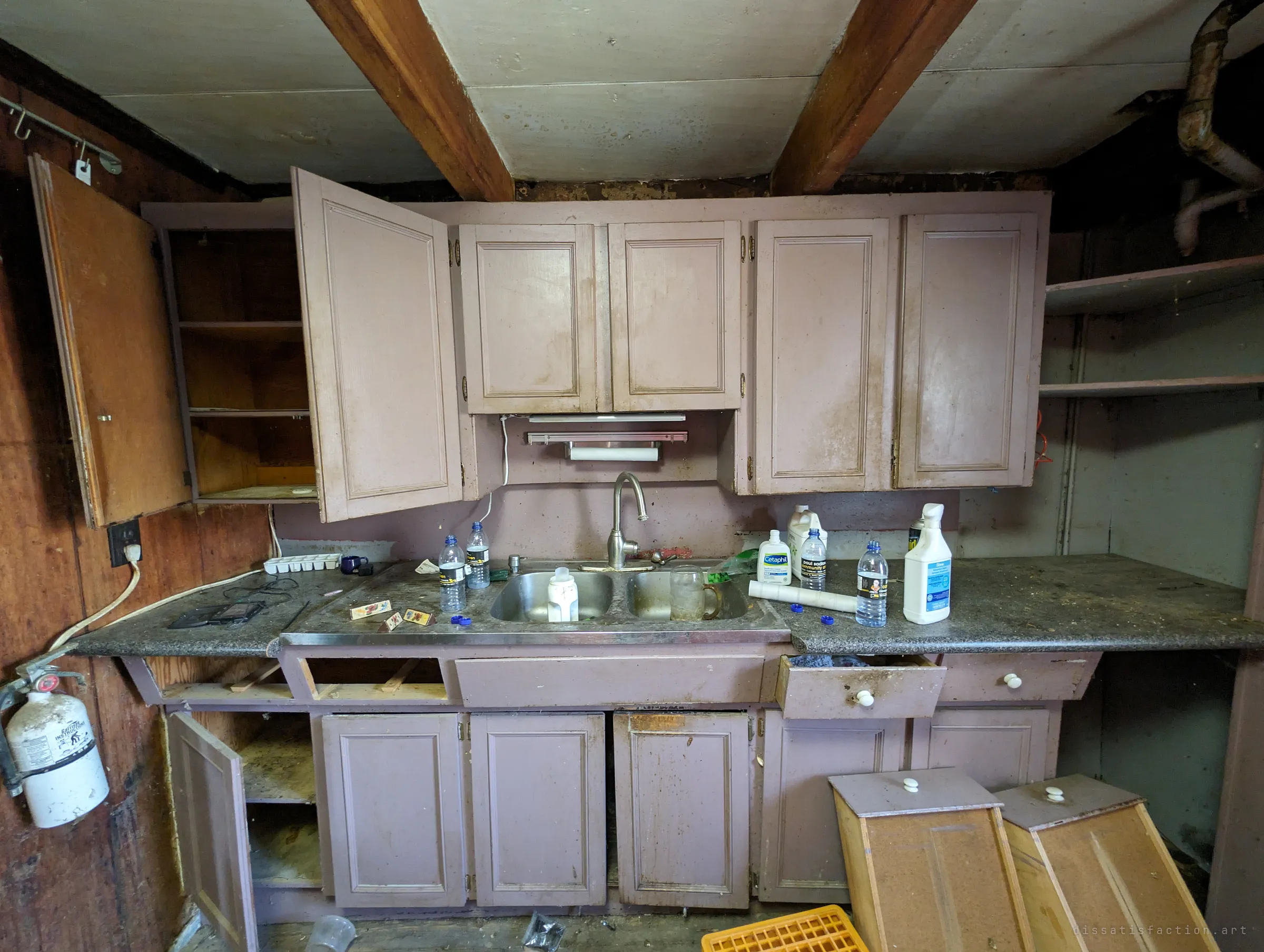 A small mauve kitchen with cabinet doors hanging open, drawers pulled out and bottles left around the sink. A fire extinguisher still hangs on the wood-panelled wall.