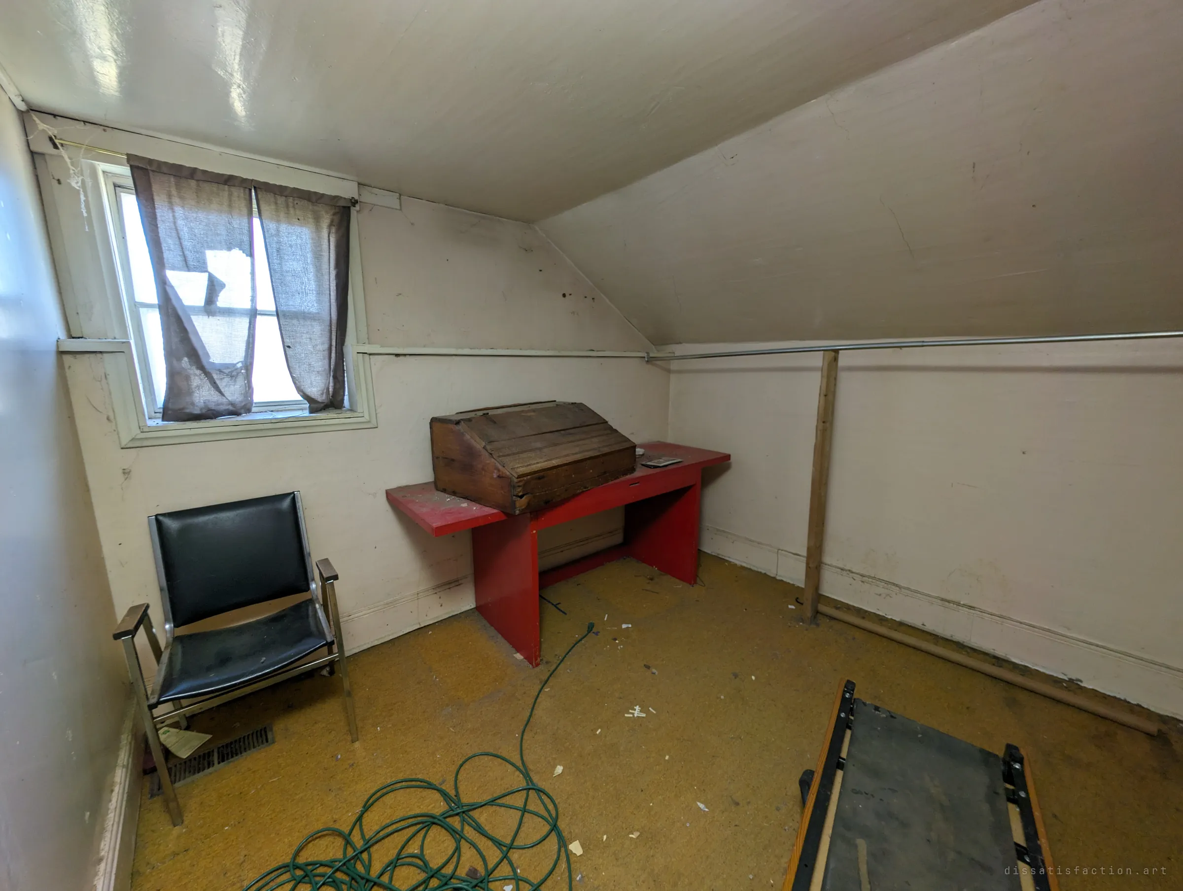 A cramped attic room with a torn curtain over the window, a black chair, a red bench holding an old wooden box and a coiled green extension cord on the mustard carpet.
