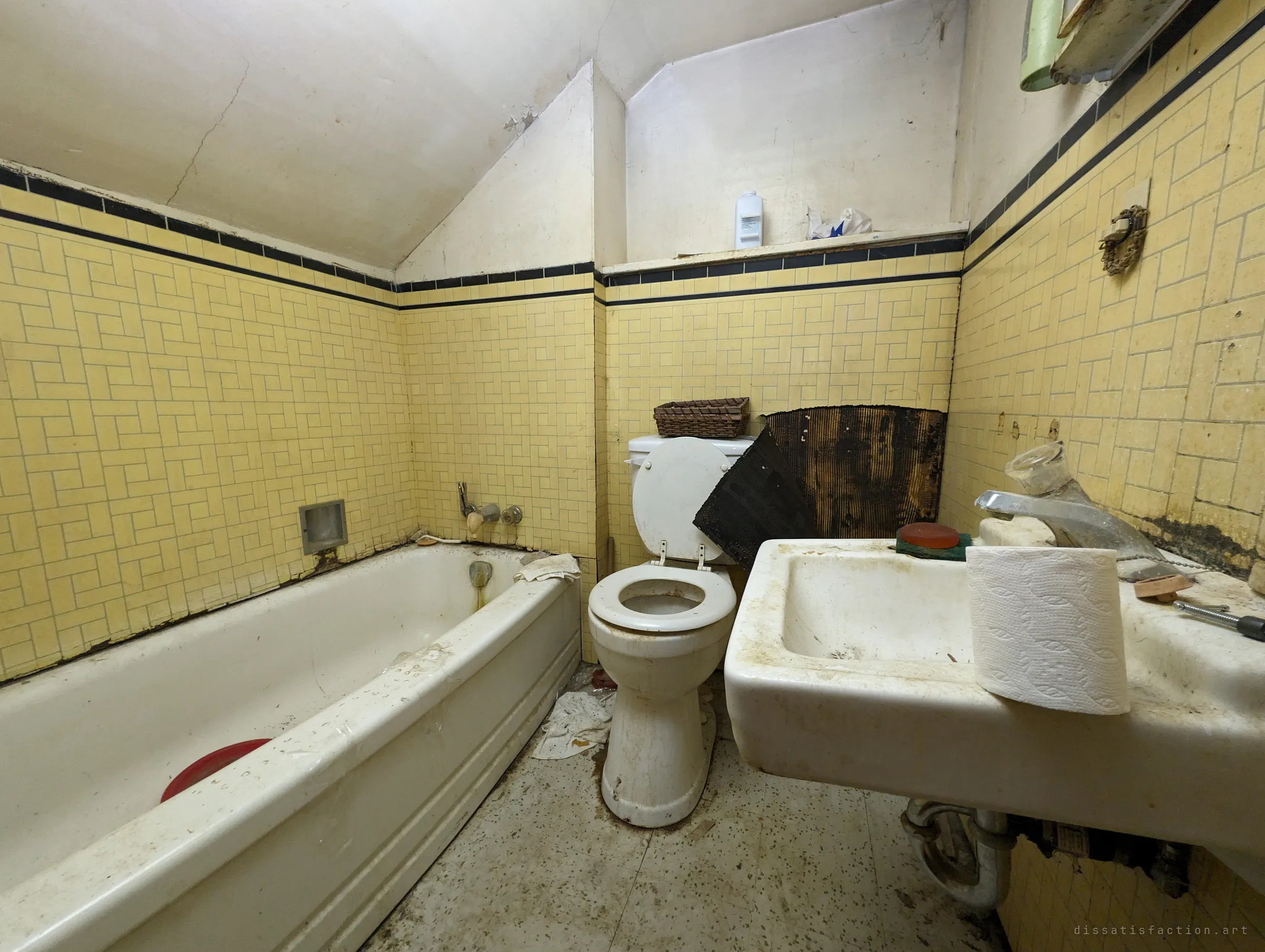 A yellow-tiled bathroom tucked under the eaves, the tub, toilet and sink stained and grimy, with a board of mould-blackened backing propped against the wall.
