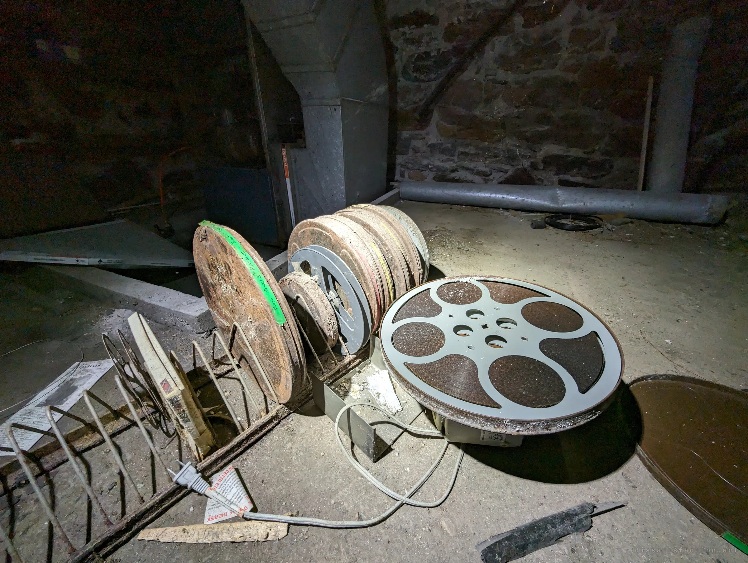Rusted film reels stacked in a wire rack on the dusty floor of a stone-walled basement, beside a large grey reel on its motor base and a galvanised furnace duct. Someone's old film collection left to corrode in the dark.