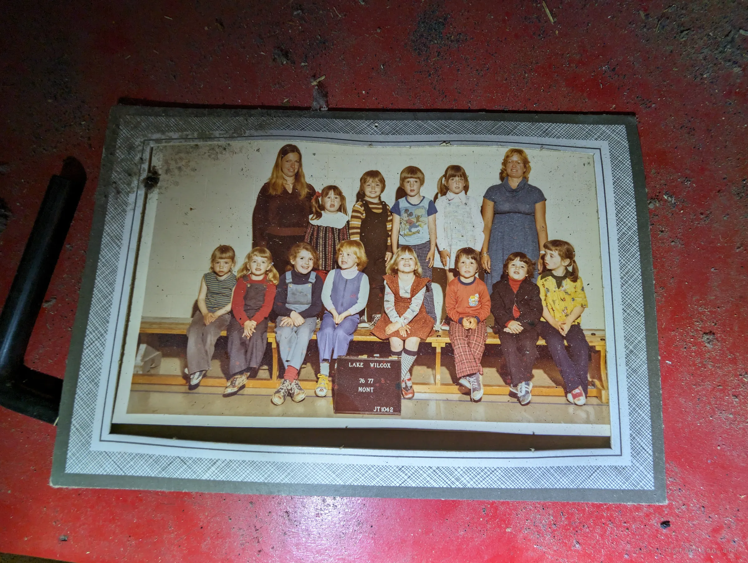 A faded 1970s school class photo in a card frame, lying on a red-painted floor. A personal keepsake left behind when the family moved out.