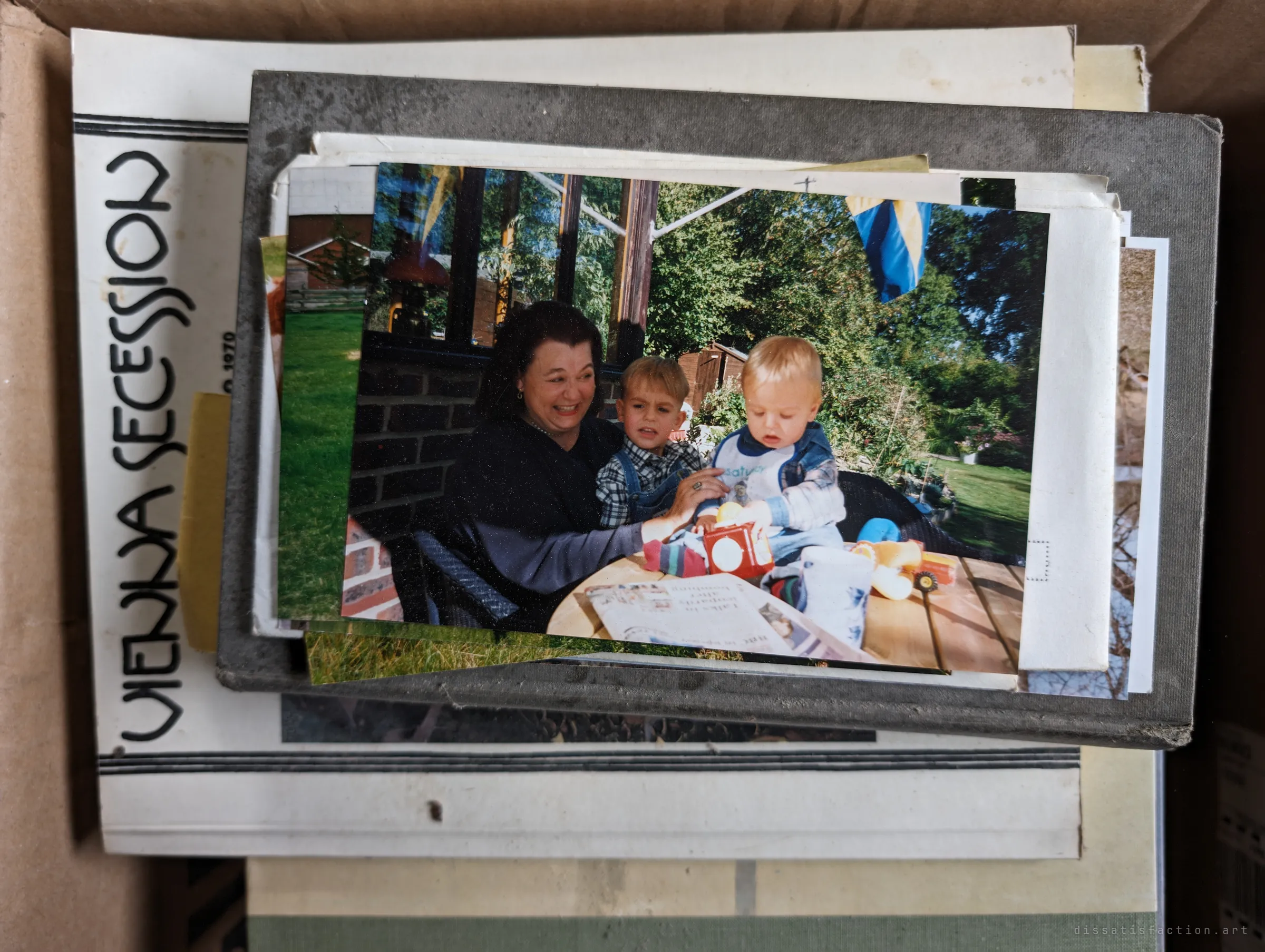 A stack of family snapshots on top of an art book in a cardboard box; the top print shows a woman laughing with two small children at a garden table. Family memories abandoned along with the house.