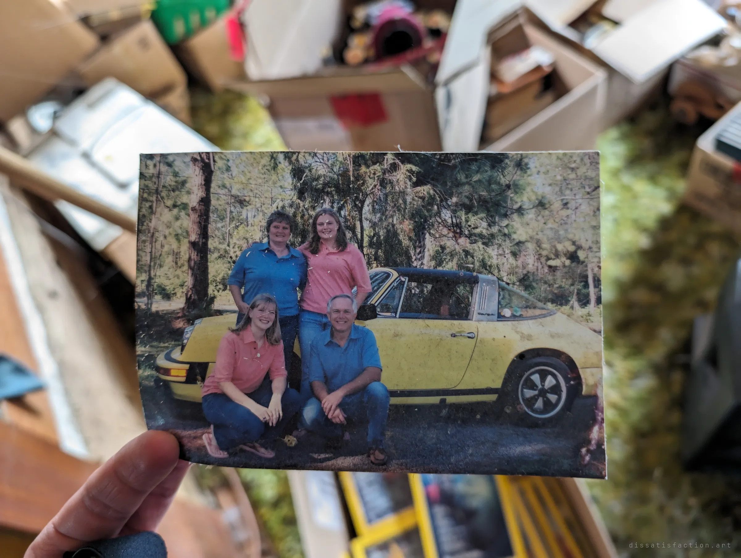 A hand holds up a found family portrait of four people posed in front of a yellow vintage convertible, over a blurred room of scattered boxes and green shag carpet. The photo shows the life that once filled the space behind it.