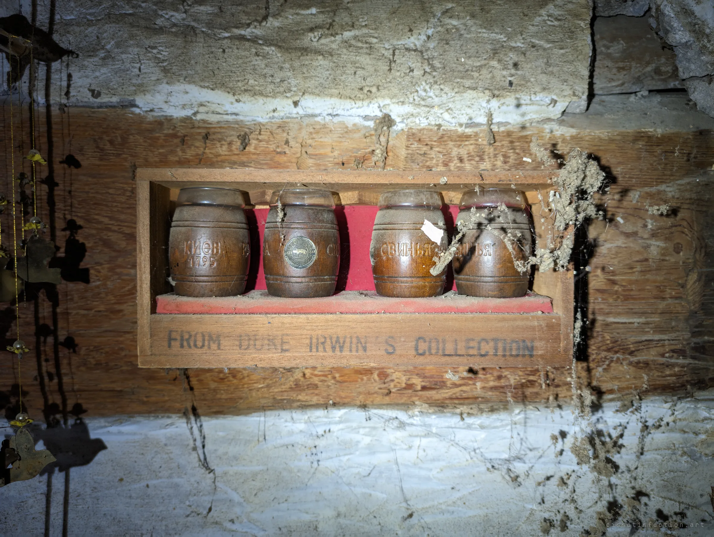 Four carved wooden barrel cups with Cyrillic lettering in a small red-lined display box on a cobwebbed wooden beam. A collector's souvenir left hanging in the dark.