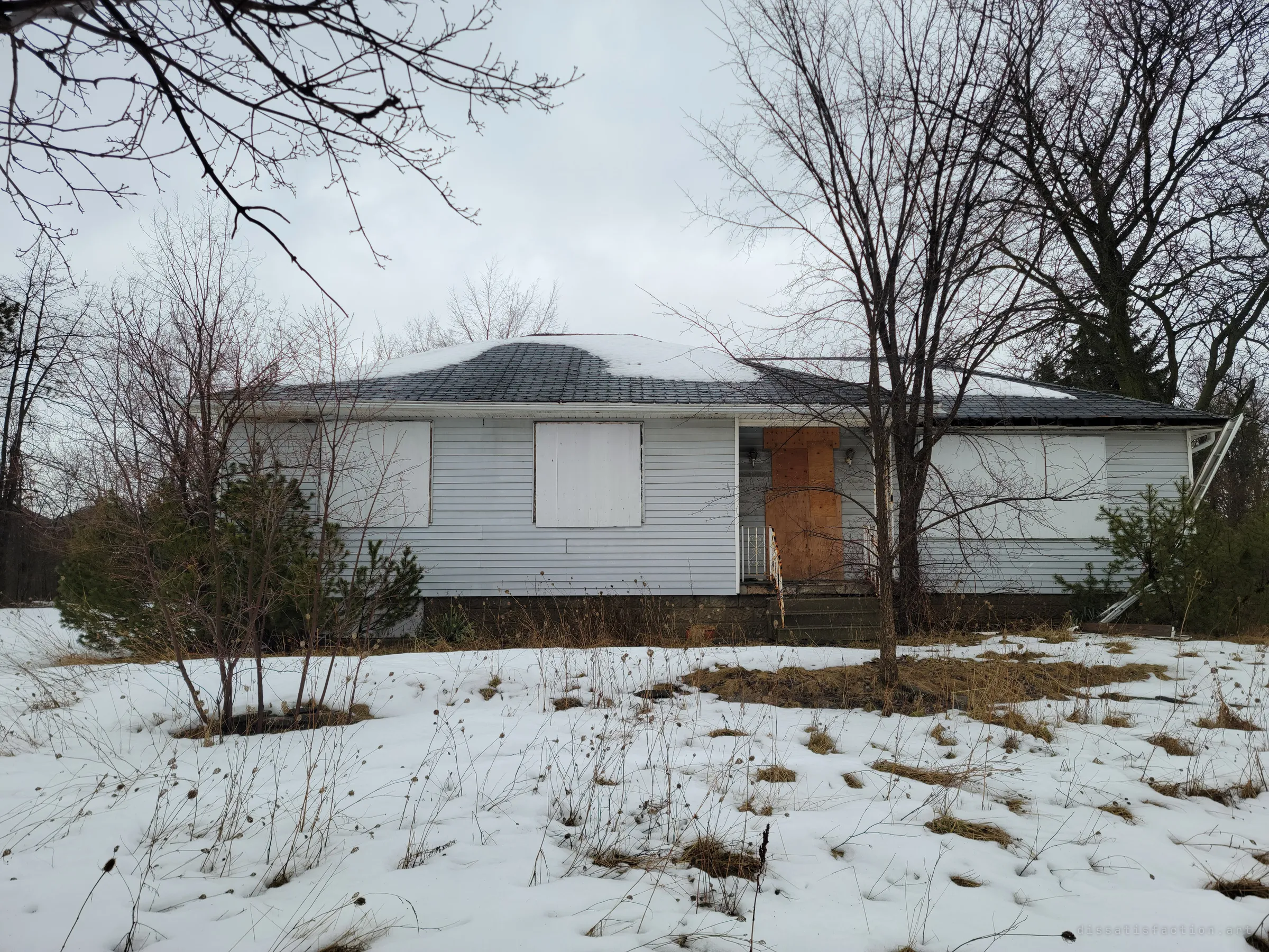 A boarded-up single-storey house with pale siding sits behind bare trees and a snowy, overgrown yard. The plywood front door and blanked windows set the tone for the abandoned home inside.