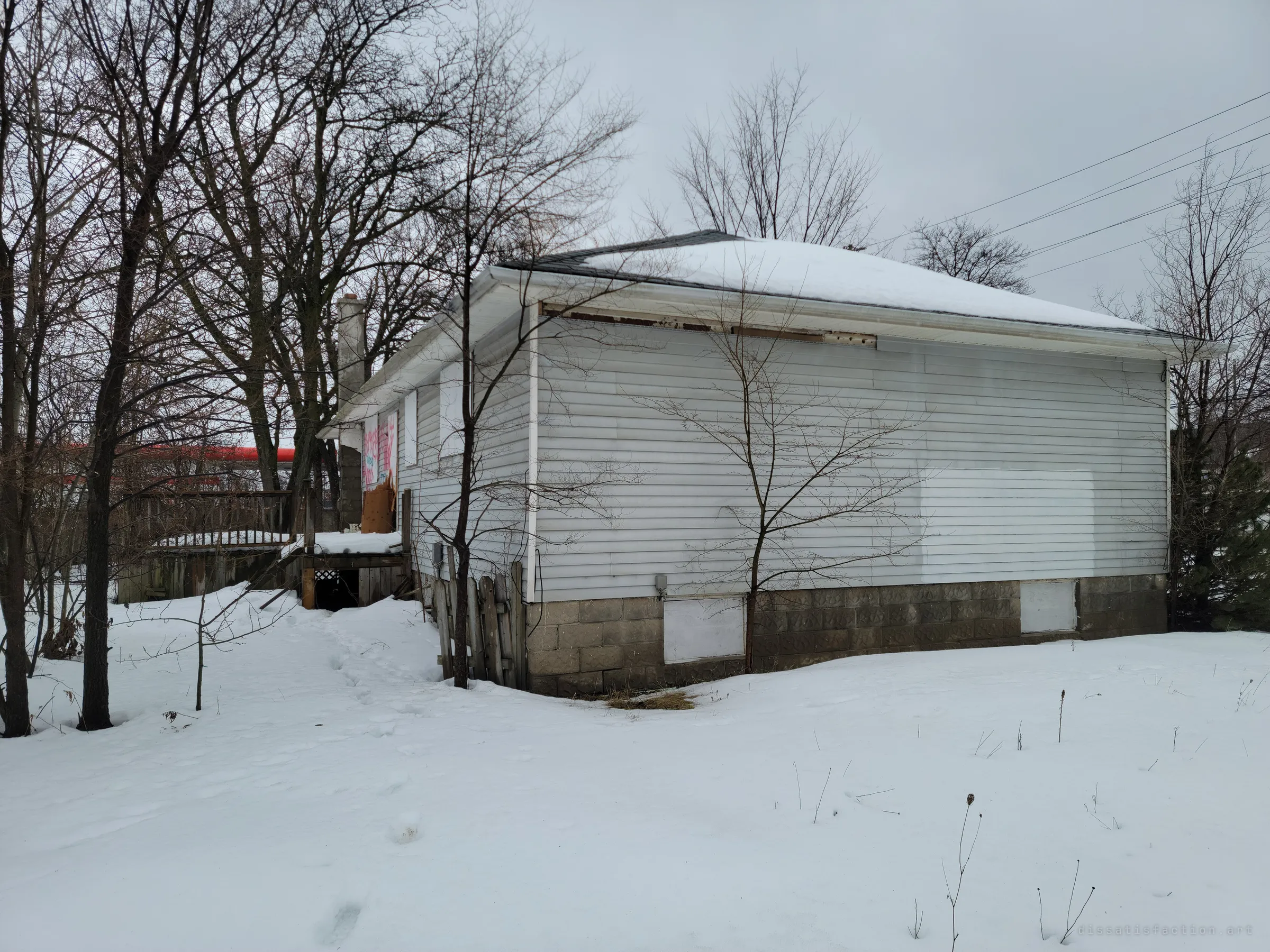 The side of the boarded house under a snow-covered hip roof, with power lines overhead and footprints in deep snow. Graffiti-covered boards are visible at the back corner.