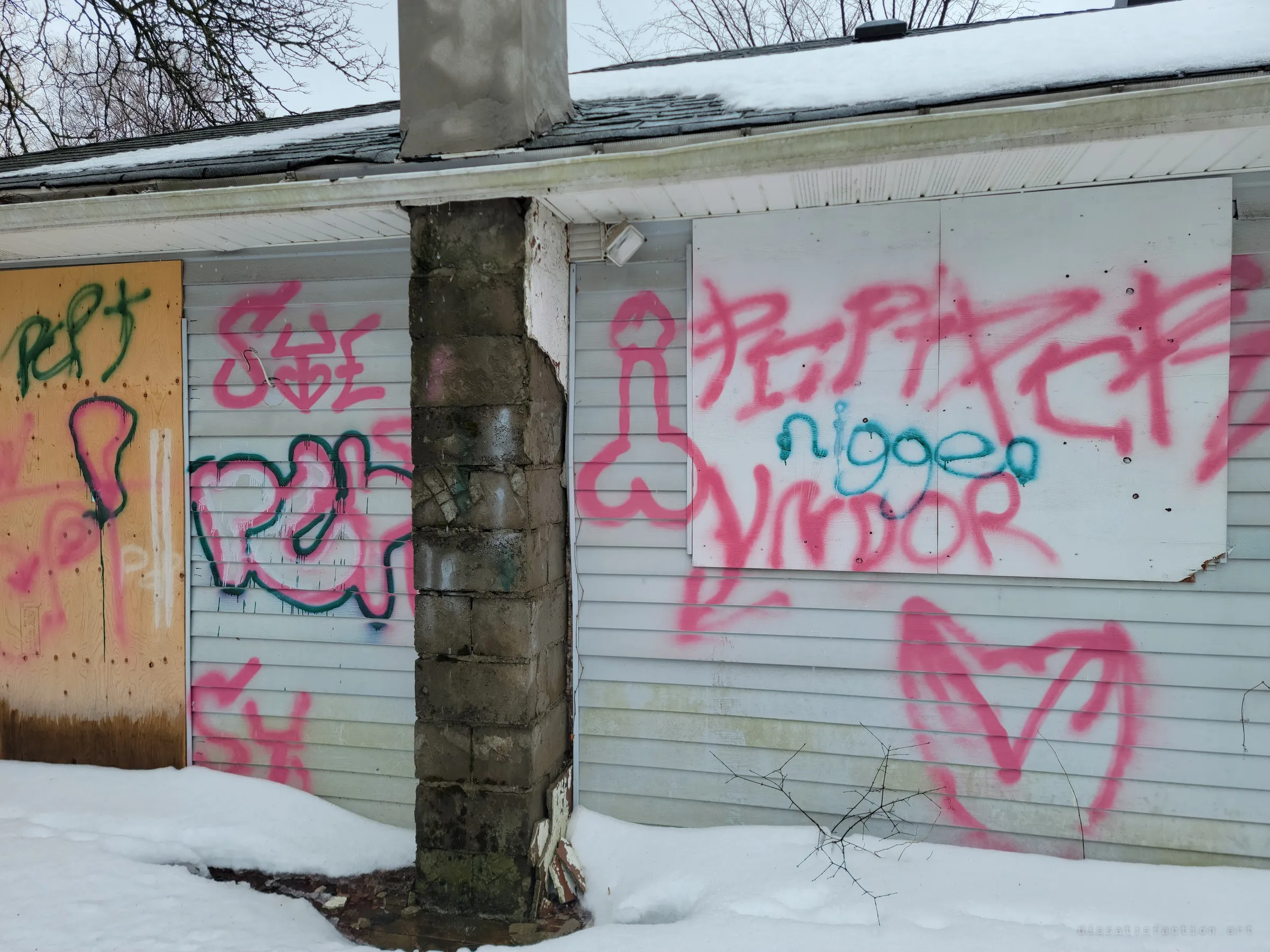 Pink and green spray-painted graffiti covers the siding and boarded windows on either side of a crumbling block chimney. Vandalism like this marks how long the house has sat open.
