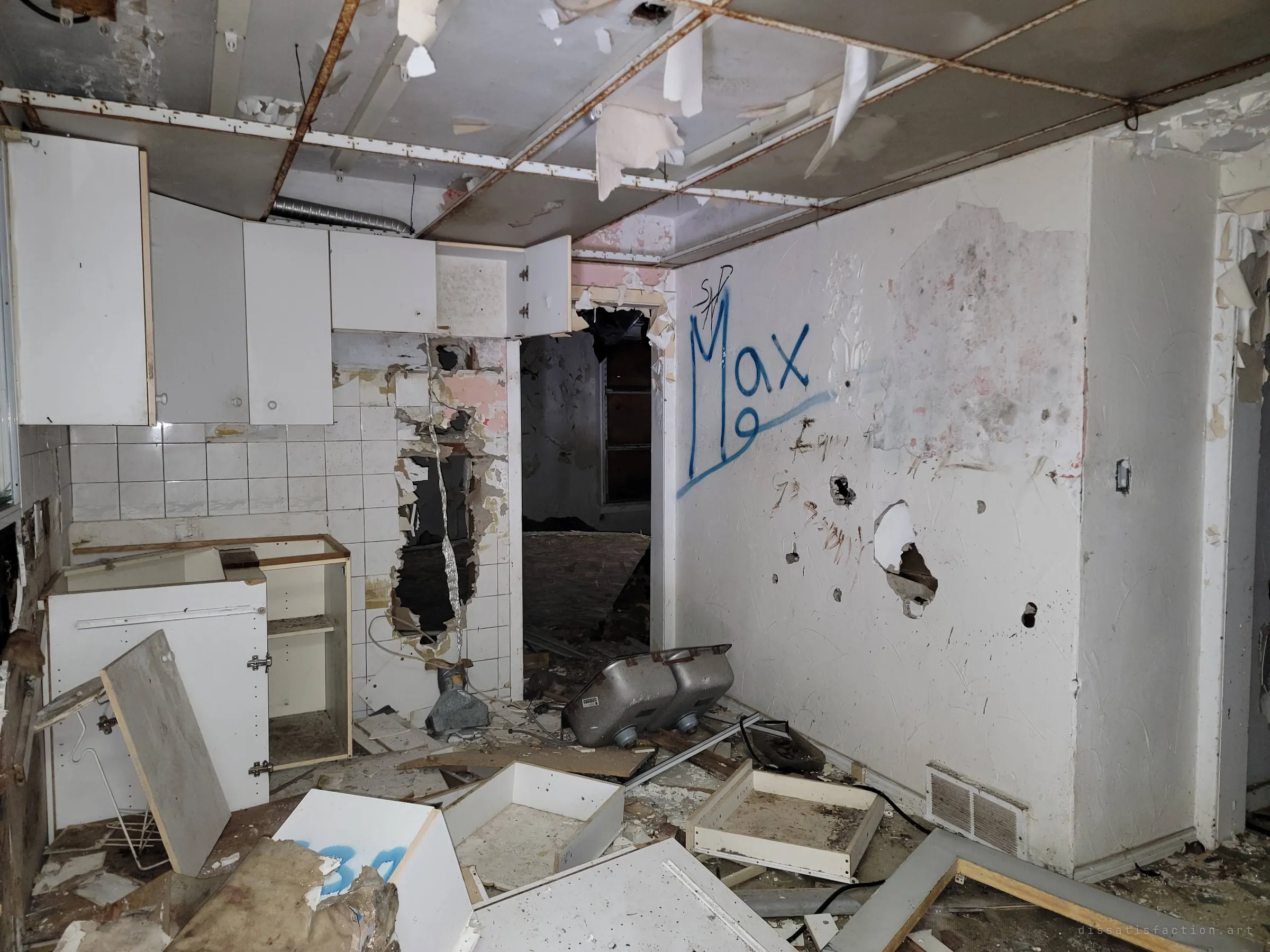 A gutted kitchen with torn-out cabinets, an upturned double sink and a sagging drop ceiling. A spray-painted name tag on the wall and holes punched through the tile show the wreckage.