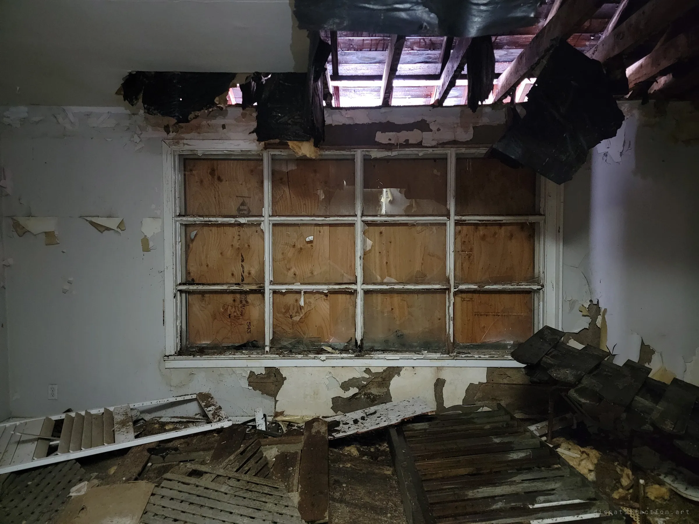 A multi-pane window boarded from outside with plywood, below a ceiling torn open to the rafters. Collapsed shutters and debris cover the floor.