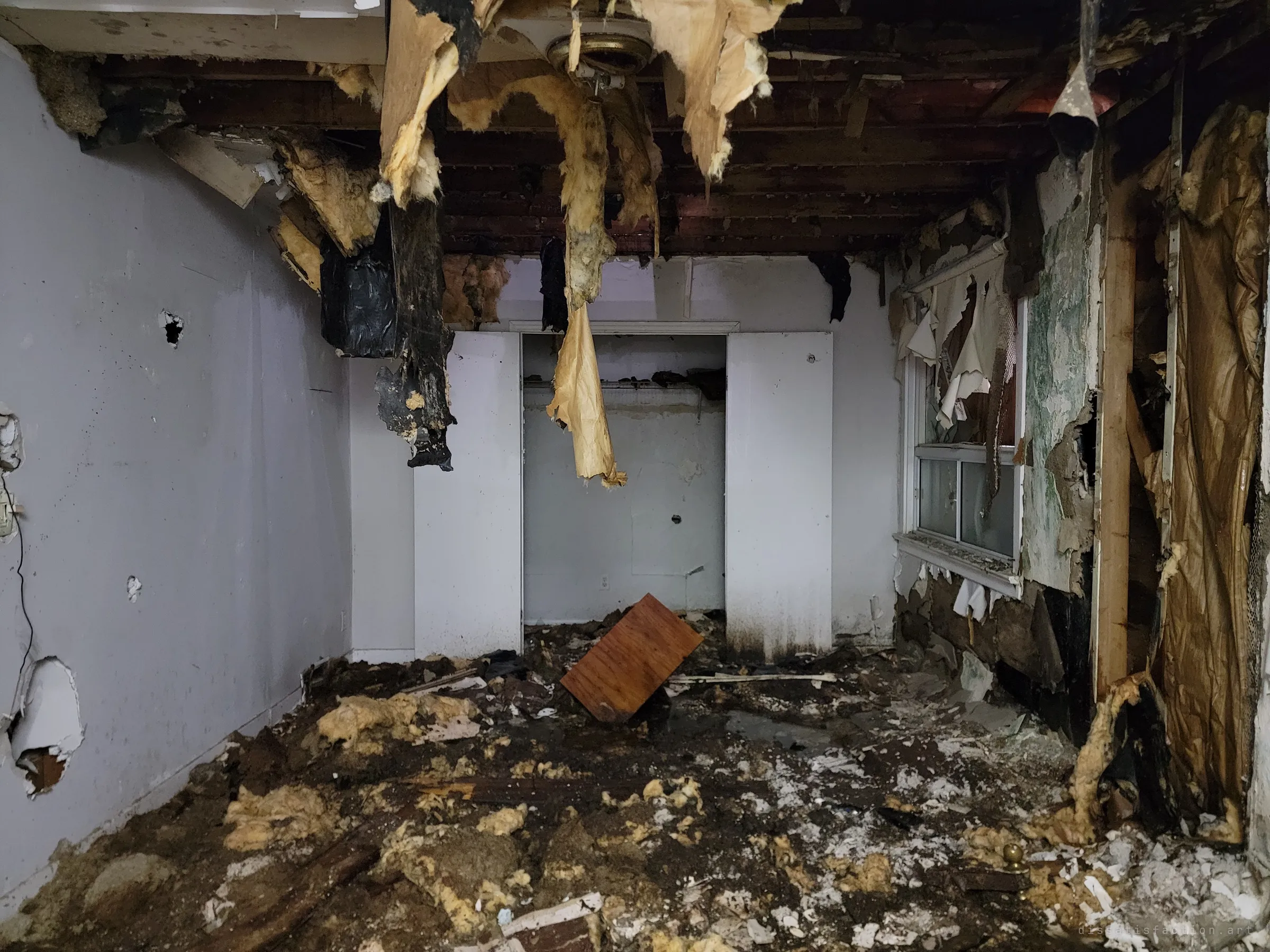 A bedroom with its ceiling ripped out, insulation and vapour barrier hanging in shreds over a floor buried in debris. An empty closet stands open at the back.