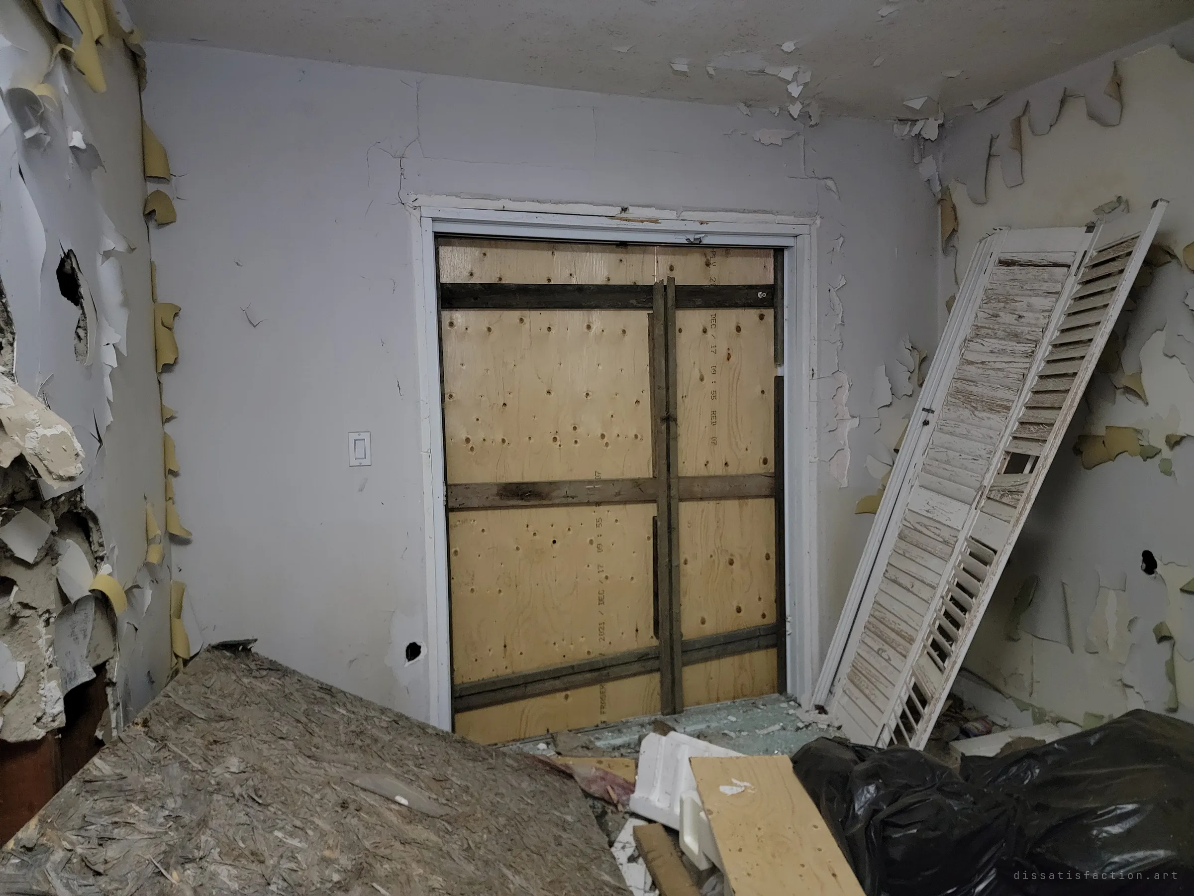 A doorway sealed with plywood and cross-battens, beside a shutter leaning against flaking walls. The boarding shows how firmly the house was closed up.