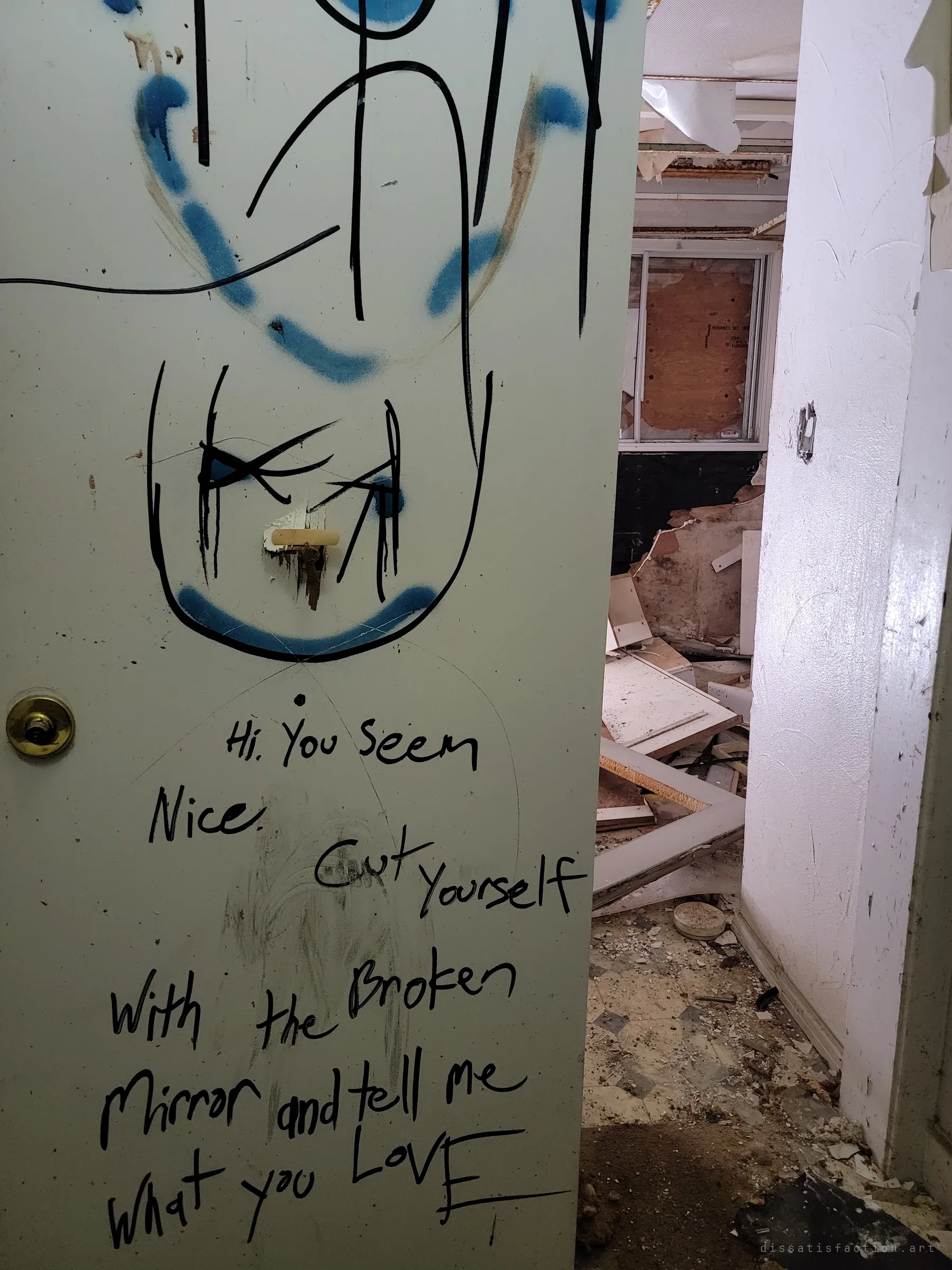 An open door covered in marker and blue spray-paint graffiti, a crude face above a scrawled taunting message, with a wrecked room of torn ceiling and a boarded window beyond.