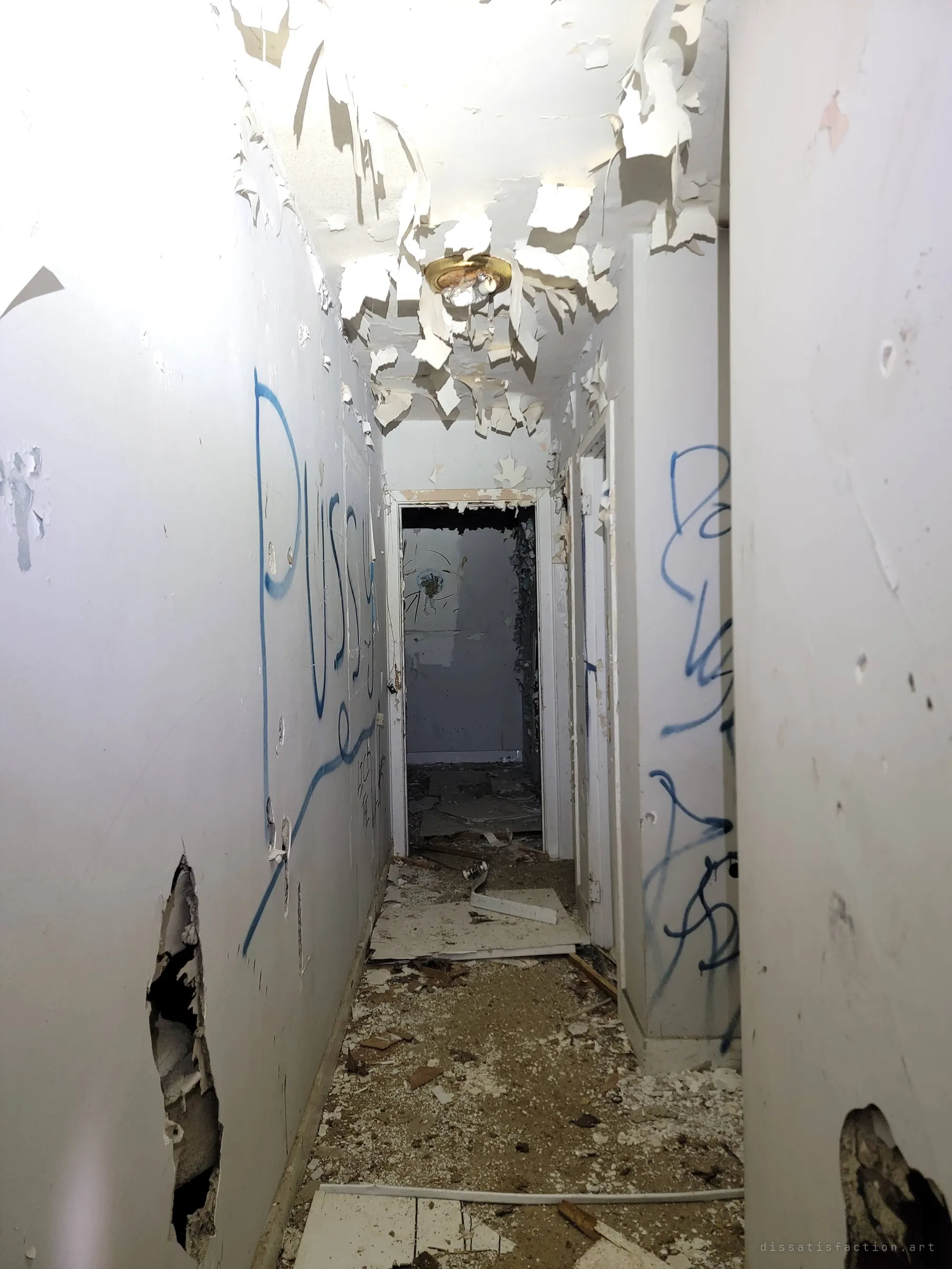 A narrow hallway with ceiling paint peeling off in curls, blue spray-paint scrawls on both walls, punched holes and debris leading to a dark doorway.