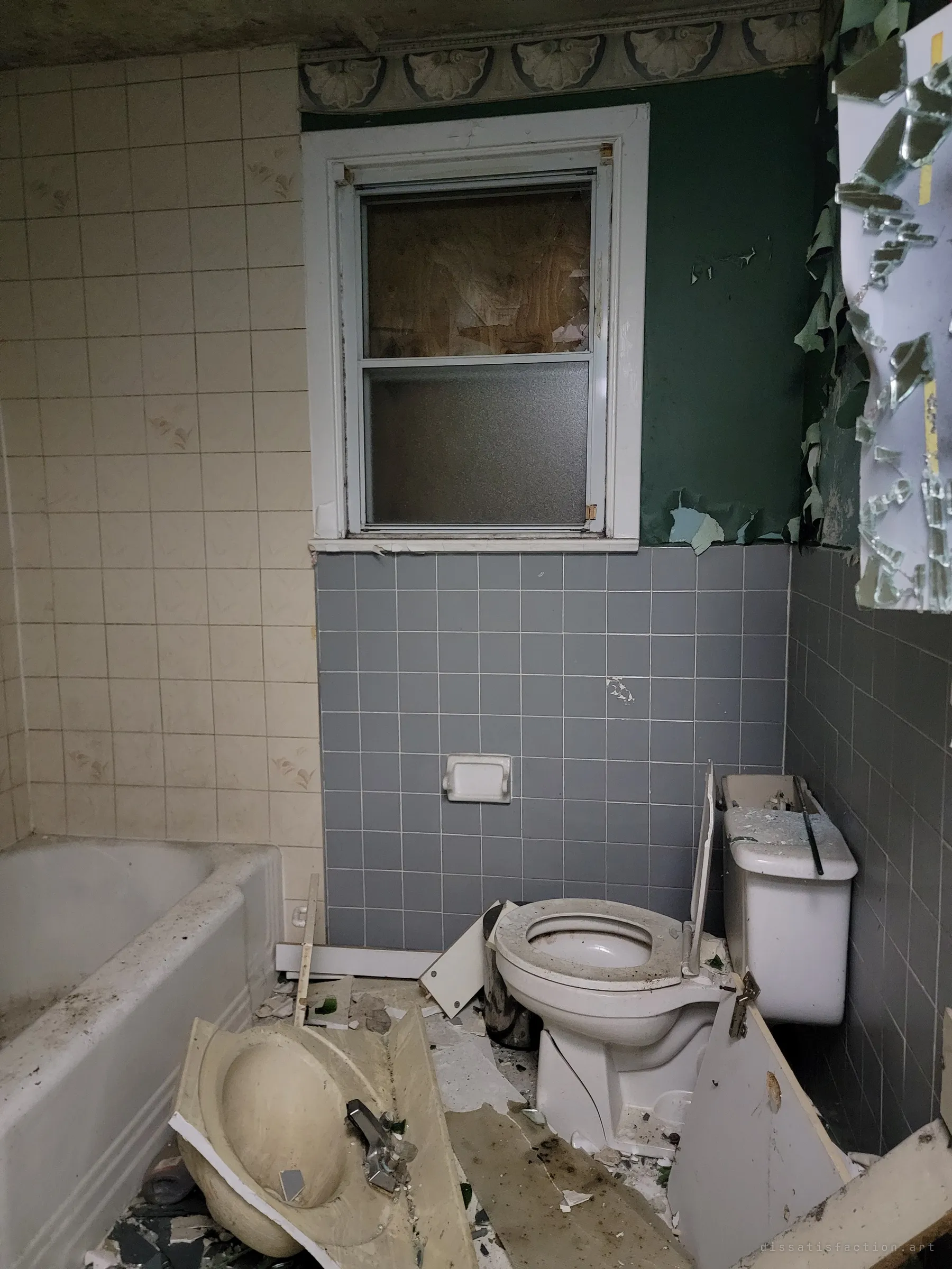 A wrecked bathroom with grey tiles and green walls, the sink ripped out and lying on the floor beside a smashed toilet tank, a shattered mirror and a boarded window.