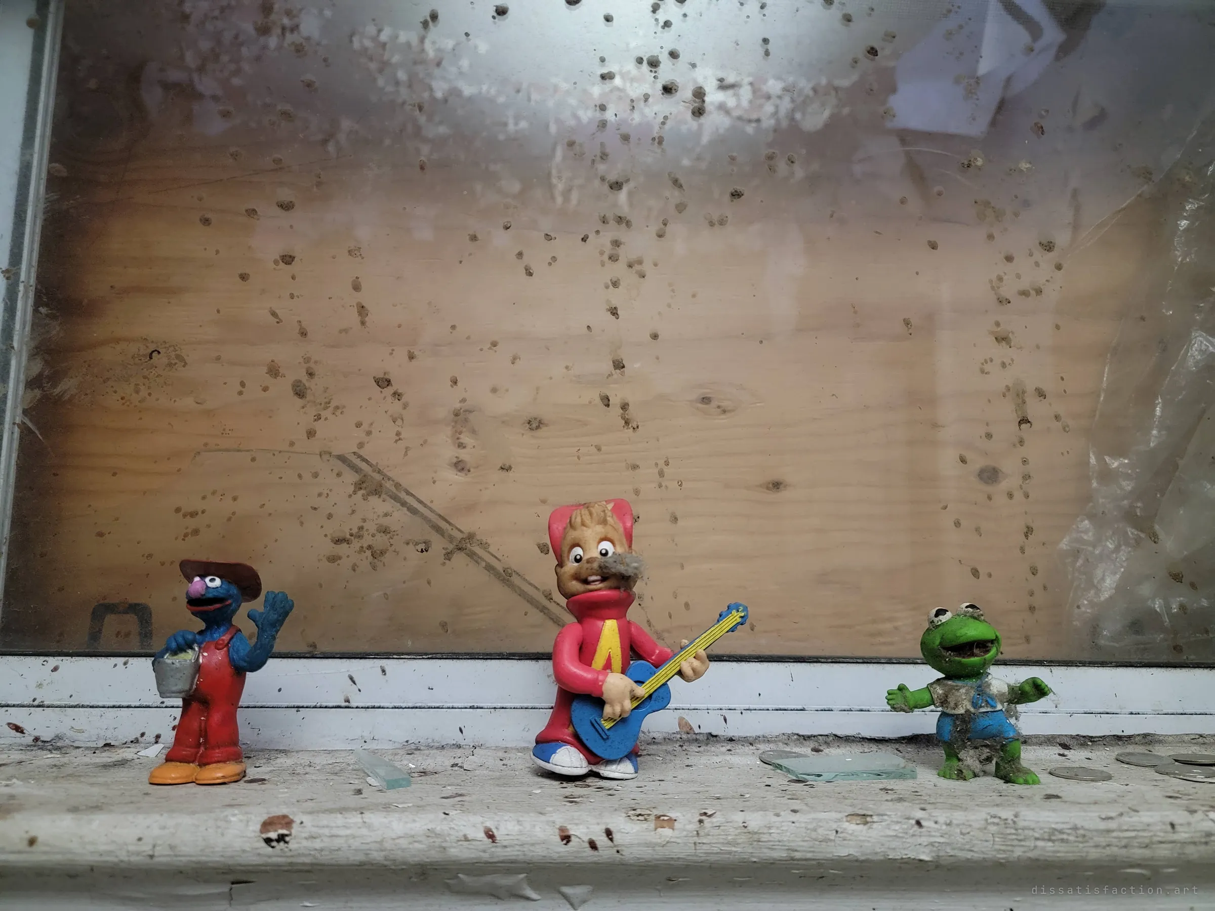 Three dusty old cartoon figurines, one playing a guitar, lined up on a windowsill in front of plywood boarding, with a few loose coins beside them.