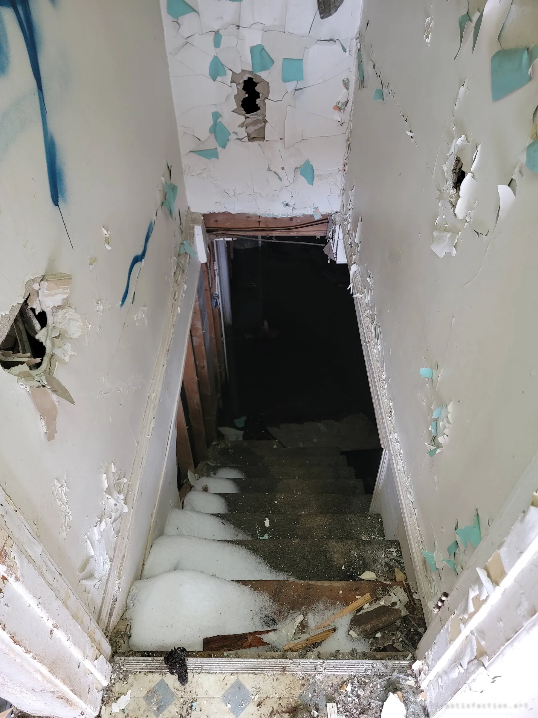 Looking down a narrow stairwell of flaking paint and broken plaster into a pitch-dark basement, the steps half-buried in white insulation foam.