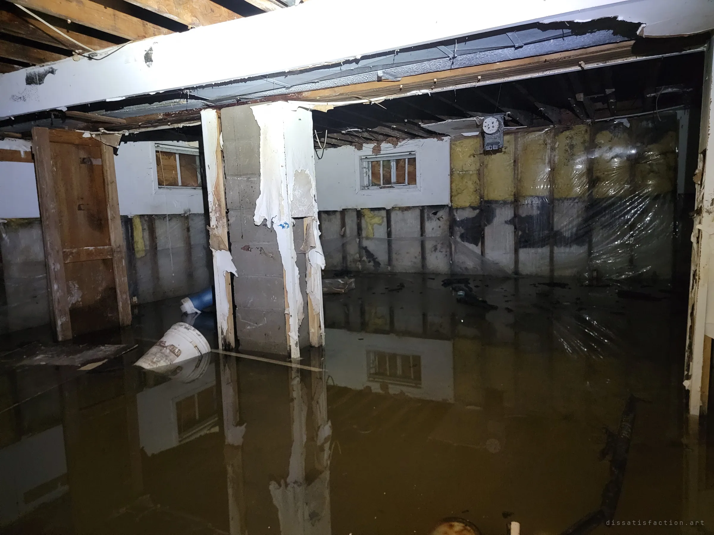 A flooded basement stripped to its studs and block walls, murky water reflecting the boarded windows, a floating bucket and a gutted concrete pillar.
