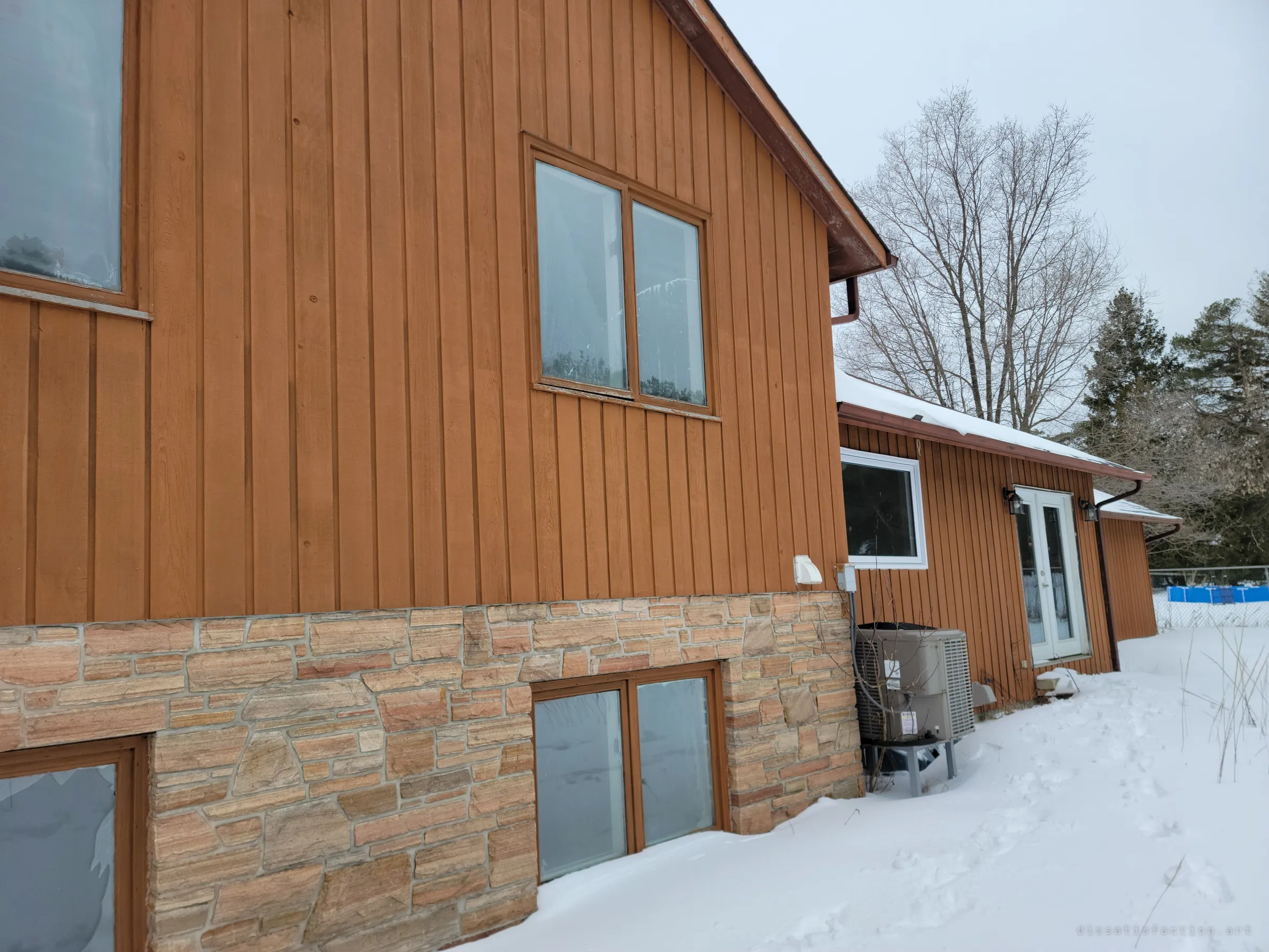 Orange board-and-batten siding over a stone foundation, with frosted windows, patio doors and an air-conditioning unit half buried in snow. The house looks intact from outside despite being empty.