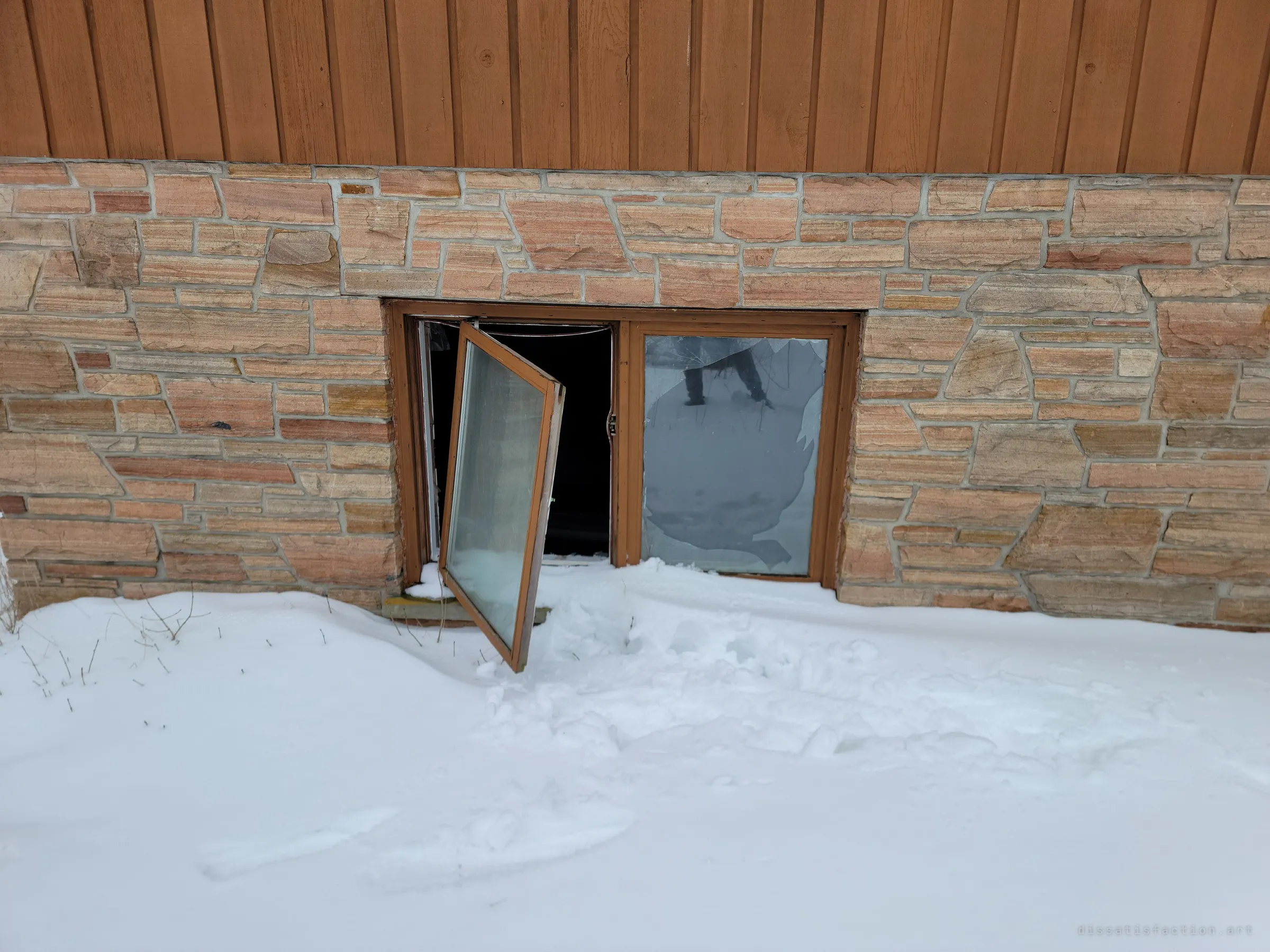 A basement window in the stone foundation hangs open on one hinge beside a smashed pane, with snow drifted against it. It shows how the house has been broken into.