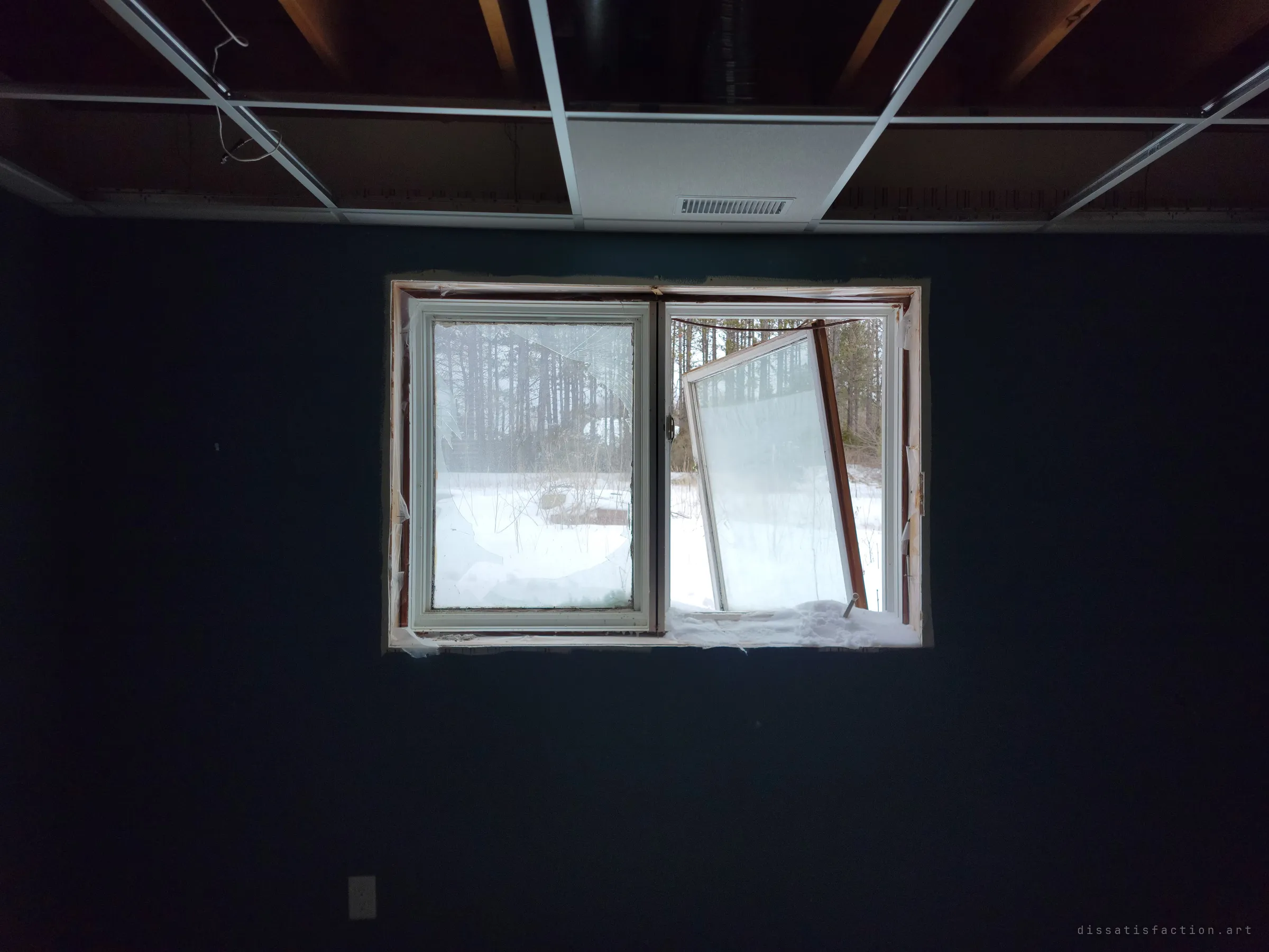 From inside a dark blue basement room, a broken window frames snow and pine trees, one sash hanging loose. The stripped drop-ceiling grid above shows the interior being gutted.