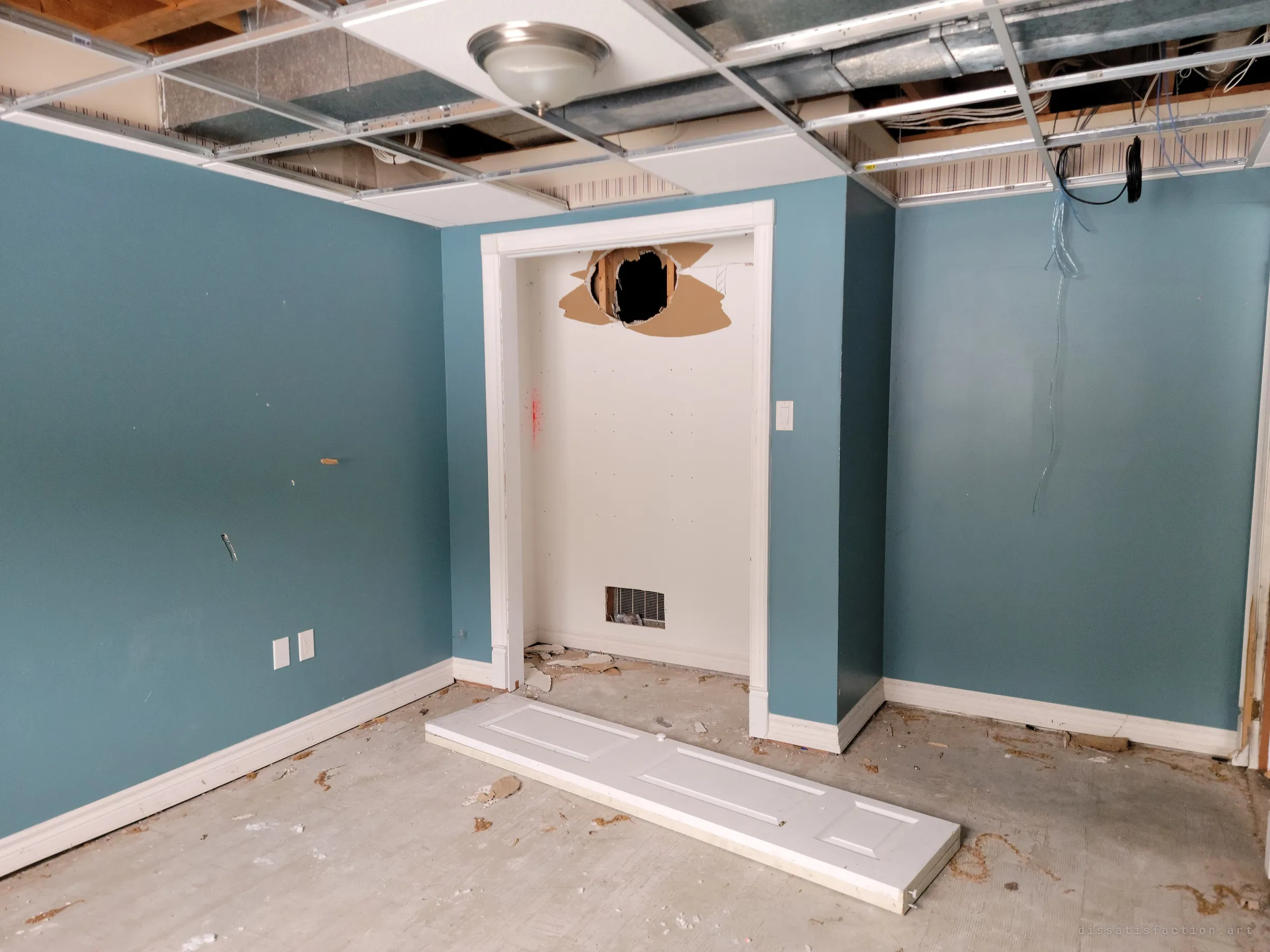 A teal basement bedroom with its closet door torn off and lying on the floor, a hole punched through the closet wall and cables dangling from the open ceiling. The damage looks deliberate.