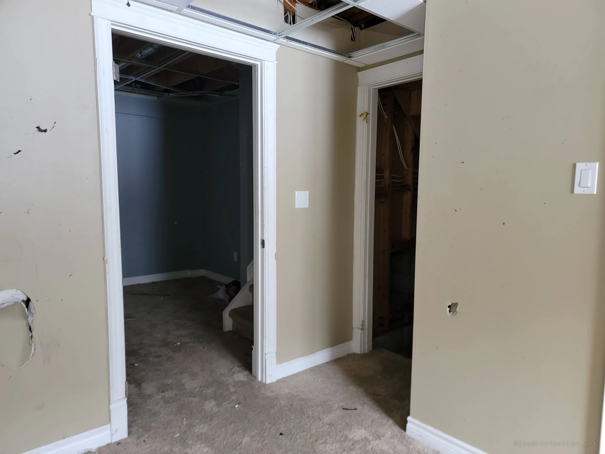 A beige basement hallway with holes in the drywall opens onto a darker room with stairs and an unfinished utility space. The carpet is littered with debris.