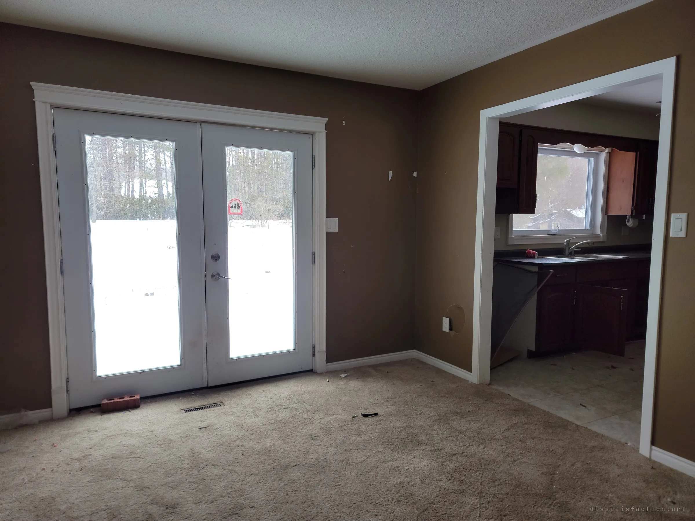 An empty brown living room with white French doors onto a snowy yard, a pet-rescue sticker still on the glass, and a kitchen with open cabinets beyond. Small traces of the family remain.