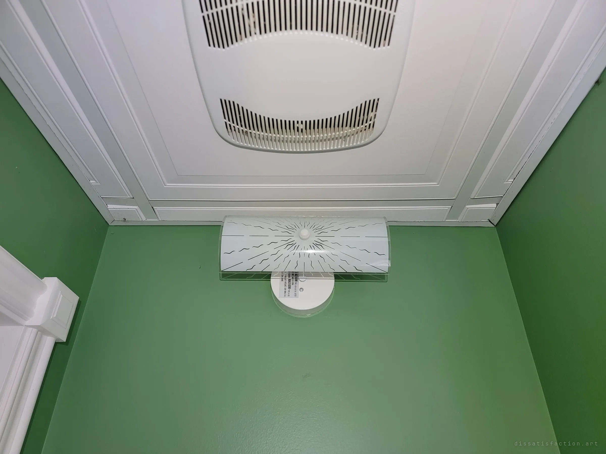 Looking up in a small green bathroom at a white ceiling fan grille and a frosted vanity light. A quiet detail that survived untouched.