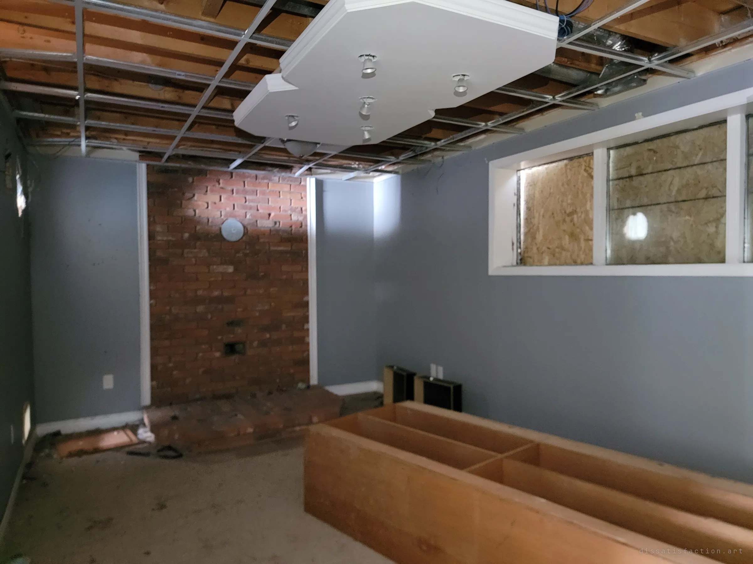 A grey basement rec room with a brick fireplace wall, boarded-up windows and a stripped ceiling grid around a hanging light panel. An empty wooden frame sits in the middle of the floor.