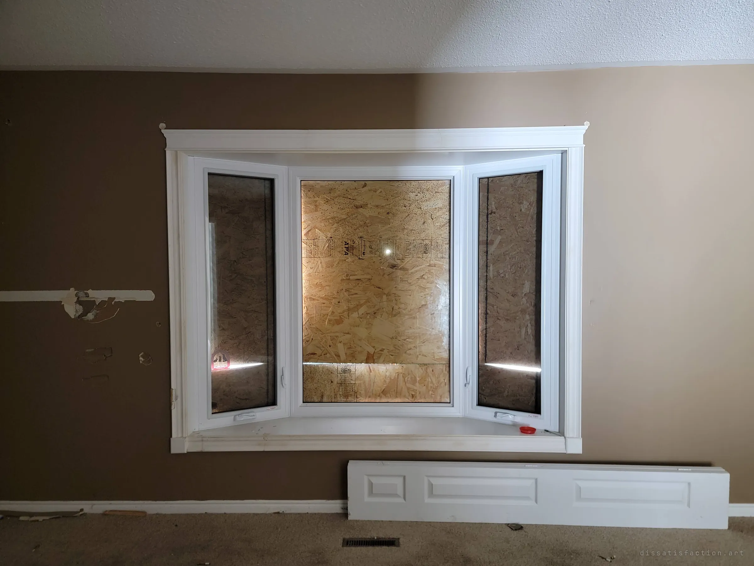 A white bay window in a brown-painted living room, every pane boarded over from outside with bare OSB, a loose panel of door trim leaning under the sill. The sealed windows mark the house as shut up and abandoned.