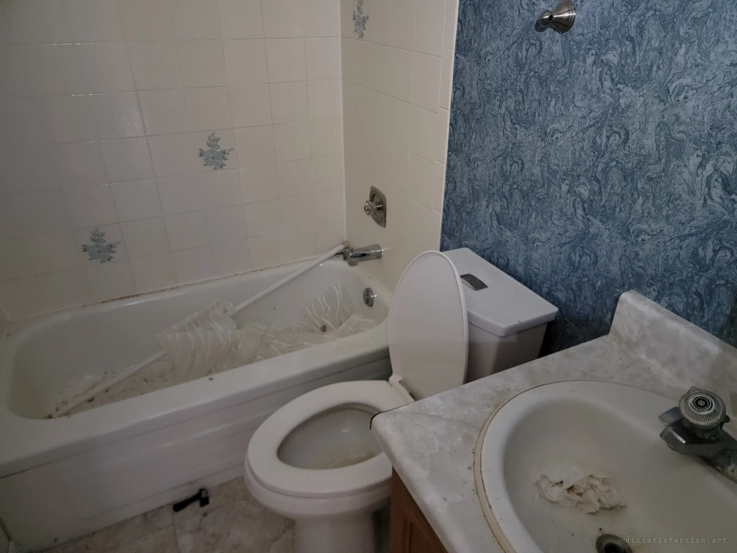 A dim bathroom with floral accent tiles and blue marbled wallpaper, a torn shower curtain and its rod collapsed into the tub beside a grimy toilet and sink. The everyday fixtures show a home left mid-life.